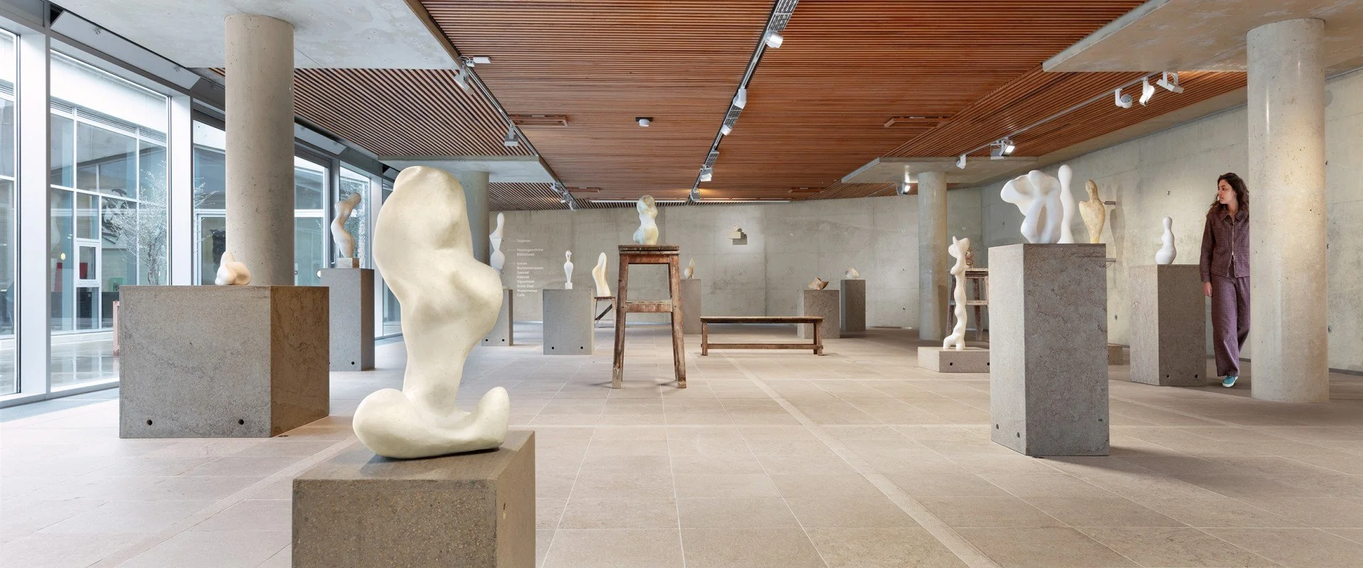Kunstgalerij met abstracte witte sculpturen op betonnen pedestals, grote ramen en houten plafond.