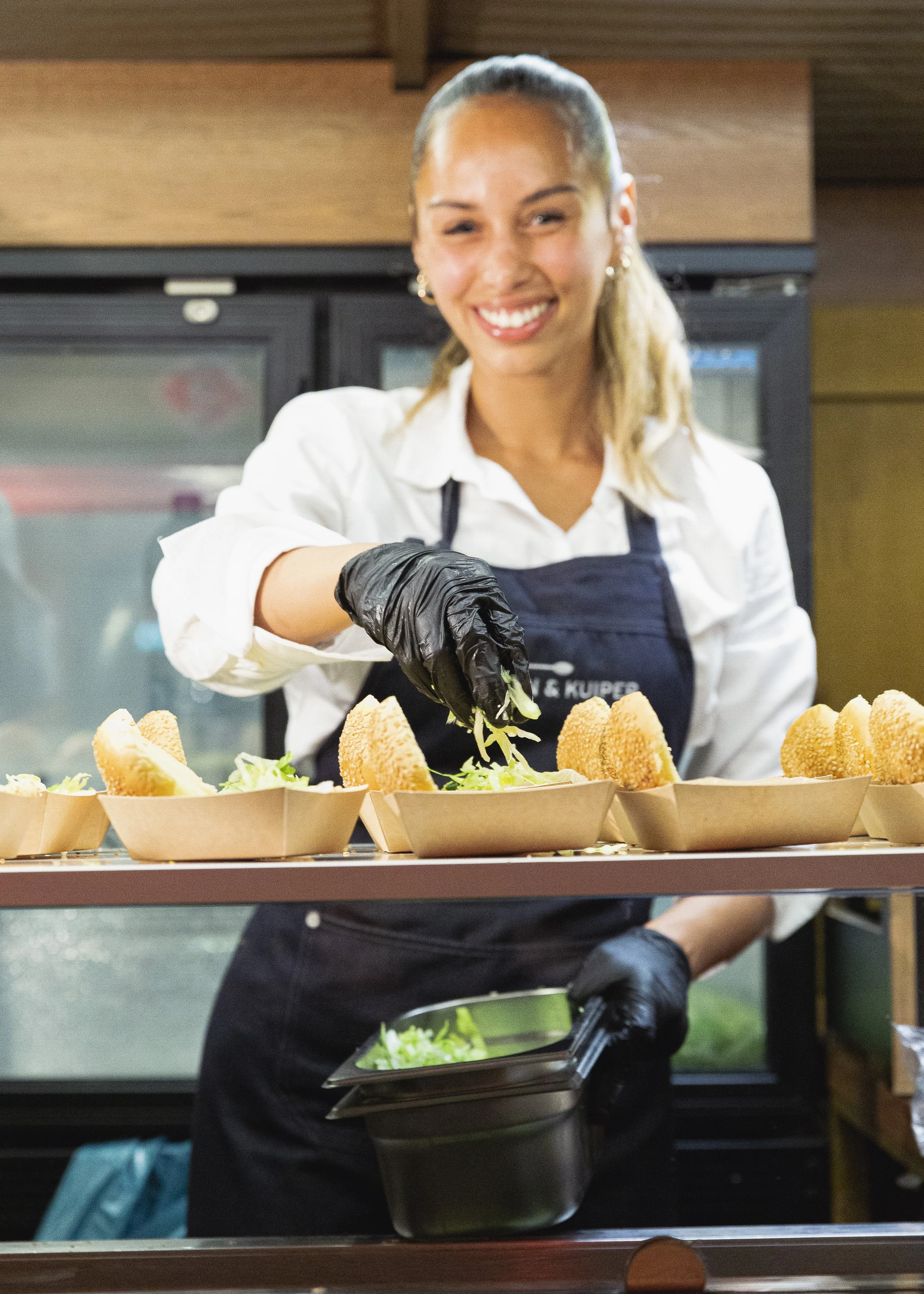 Vrouw met zwarte handschoenen die voorbereidt en serveert sandwiches met groen garnituur in een restaurantkeuken.