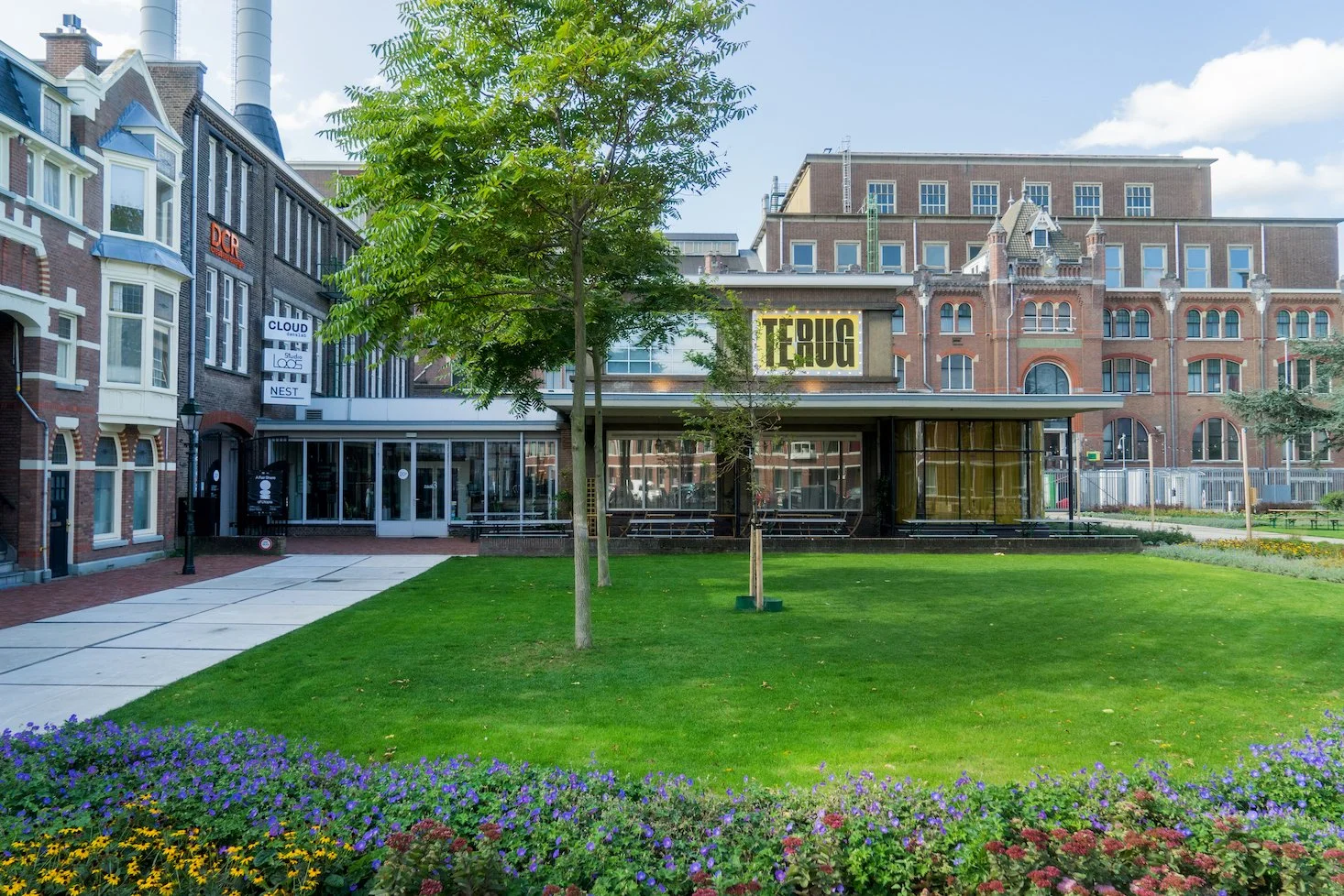 Groene tuin met bloemen en bomen voor een modern gebouw met grote ramen en een digitale scherm.beelden de woorden 'TEUG'