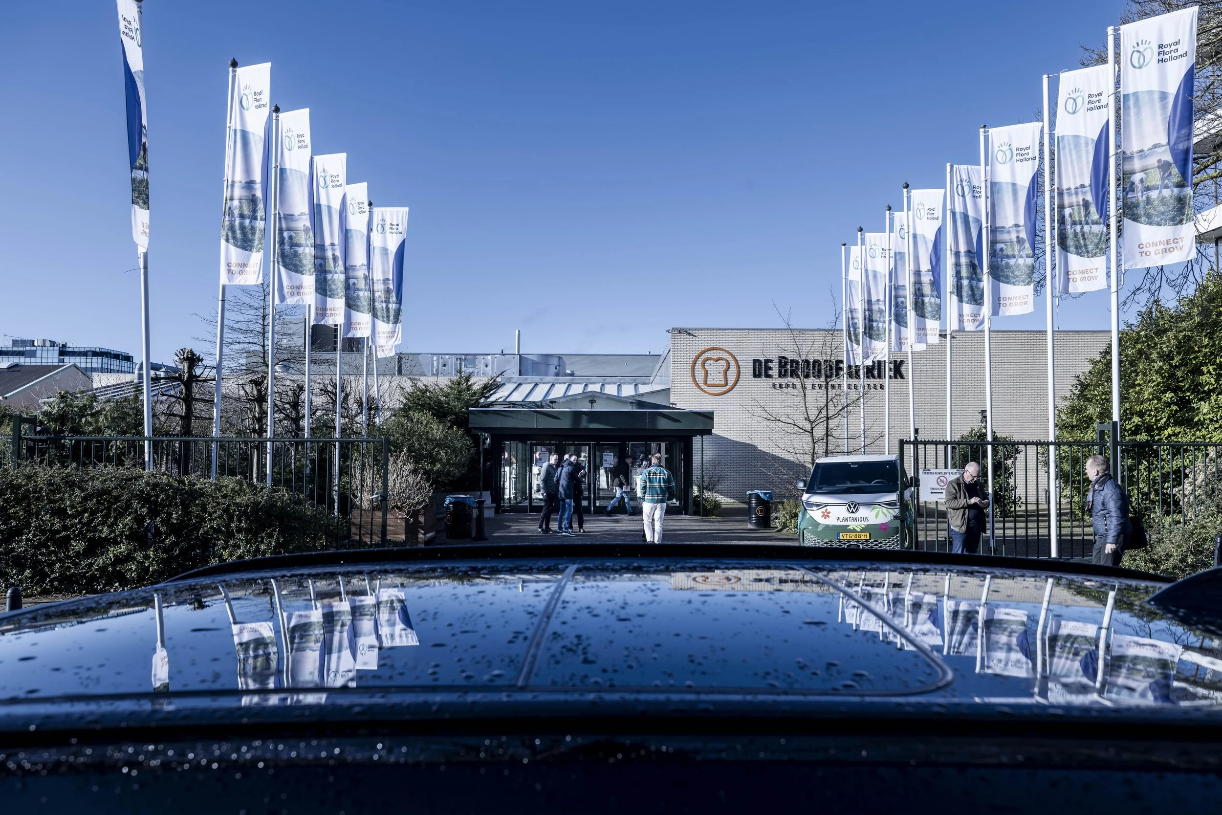 De ingang van een broodfabriek met een groep mensen die naar binnen gaat, vlaggen met het logo van Royal Flora Holland en een auto voor de ingang.