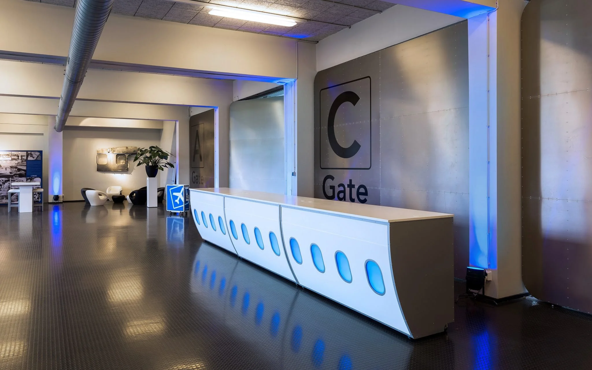 In een luchthaventerminal is een witte balie met blauwe verlichting, achter een groot metalen paneel met het woord "Gate" en een grote letter "C".