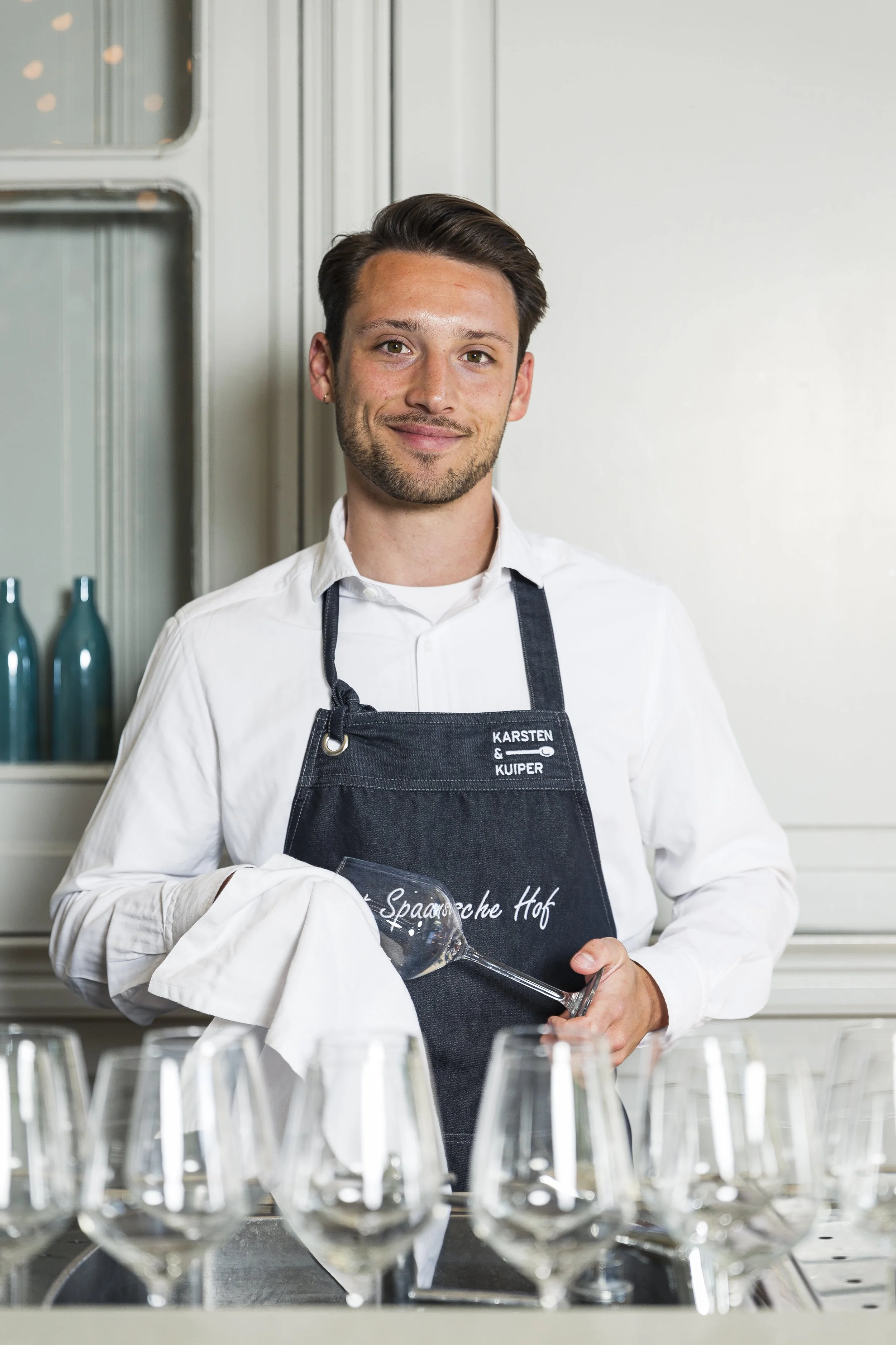 Man in witte blouse en zwart schort met druiventros en wijnglas.