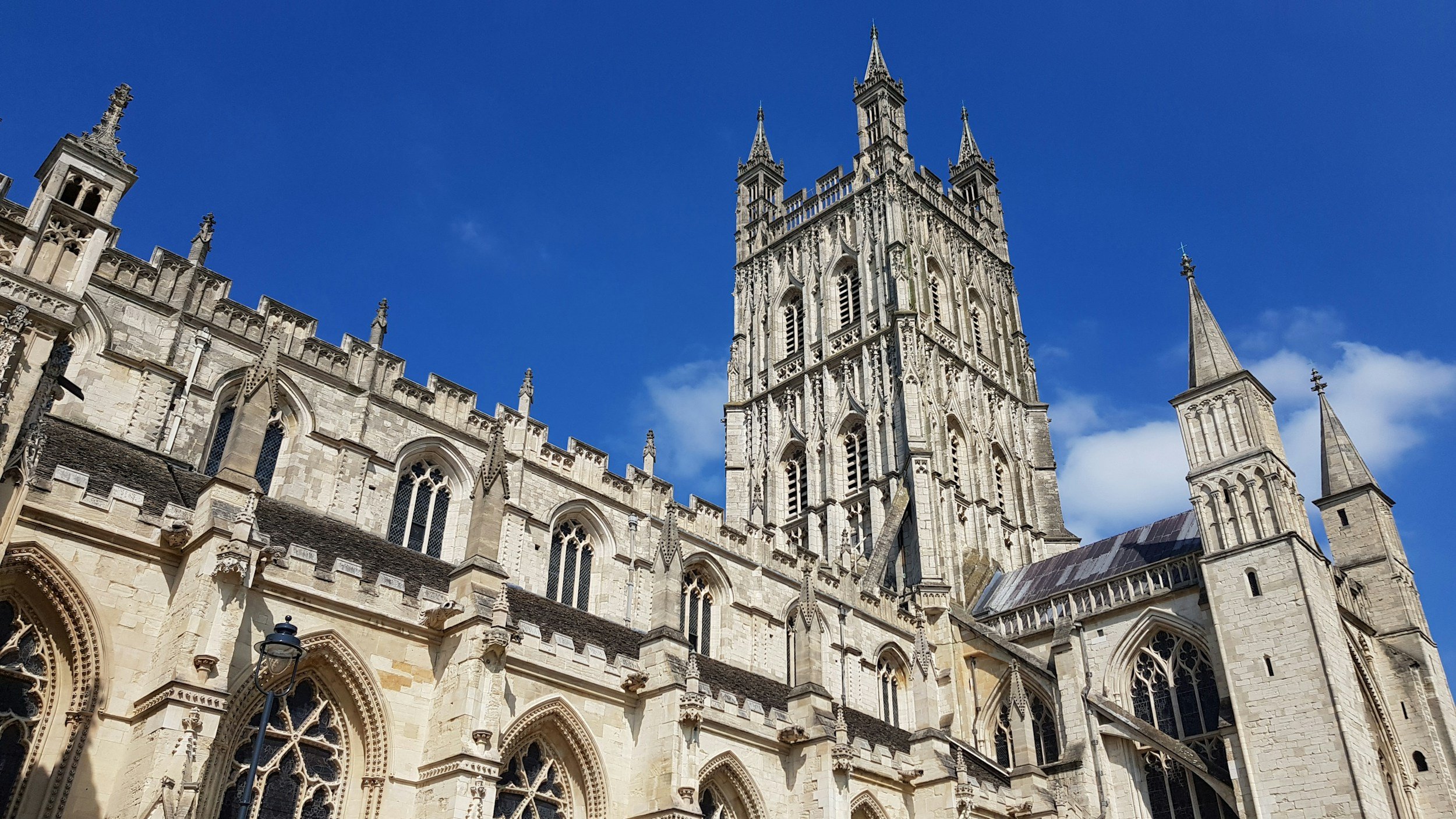A panoramic view of a historic Gothic-style cathedral with intricate stone architecture, tall spires, and stained glass windows against a bright blue sky with a few clouds.