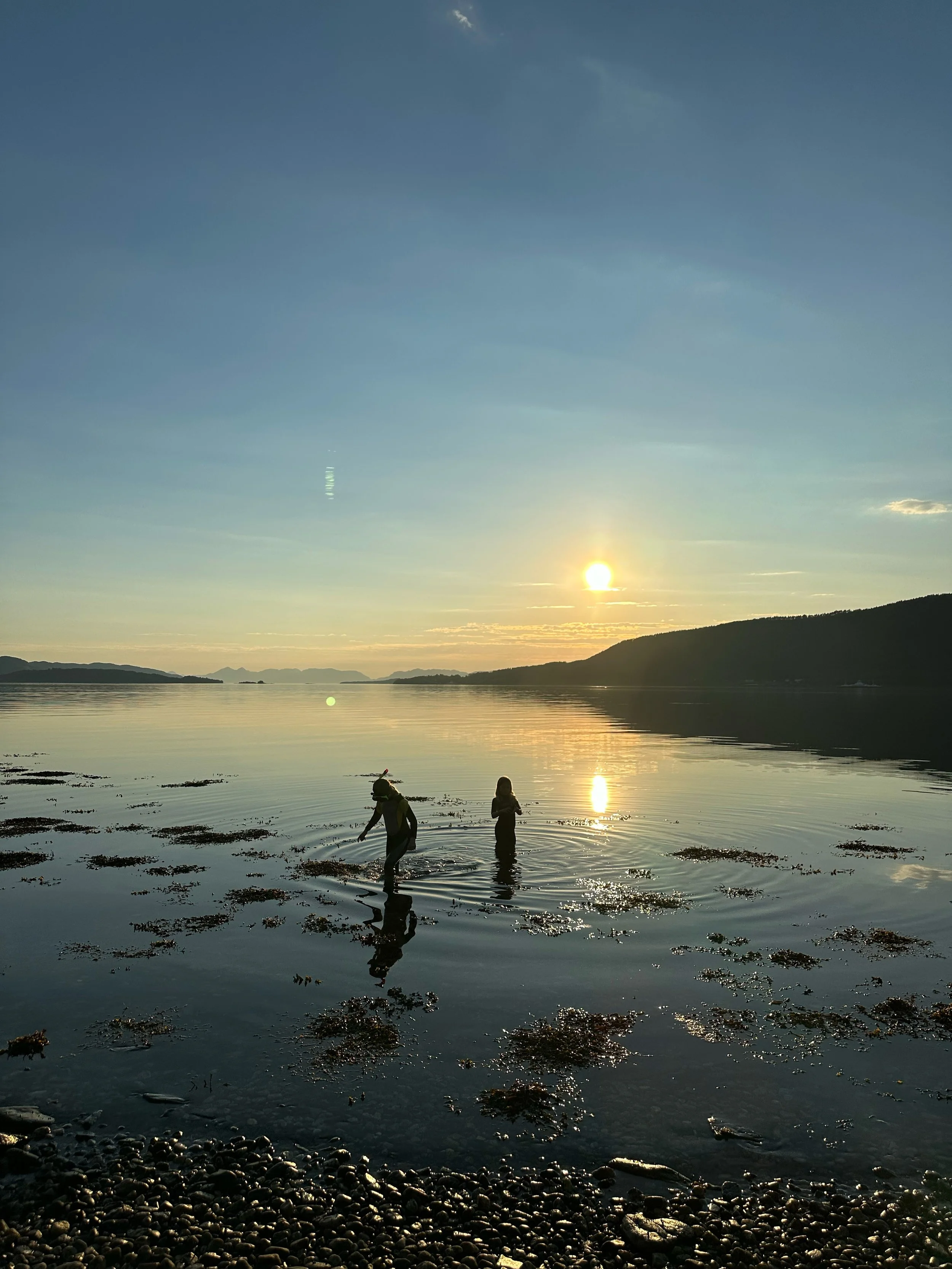 To personer som står i vannet under solnedgang, omringet av fjell og sjø, med en klar himmel og refleksjoner i vannet.