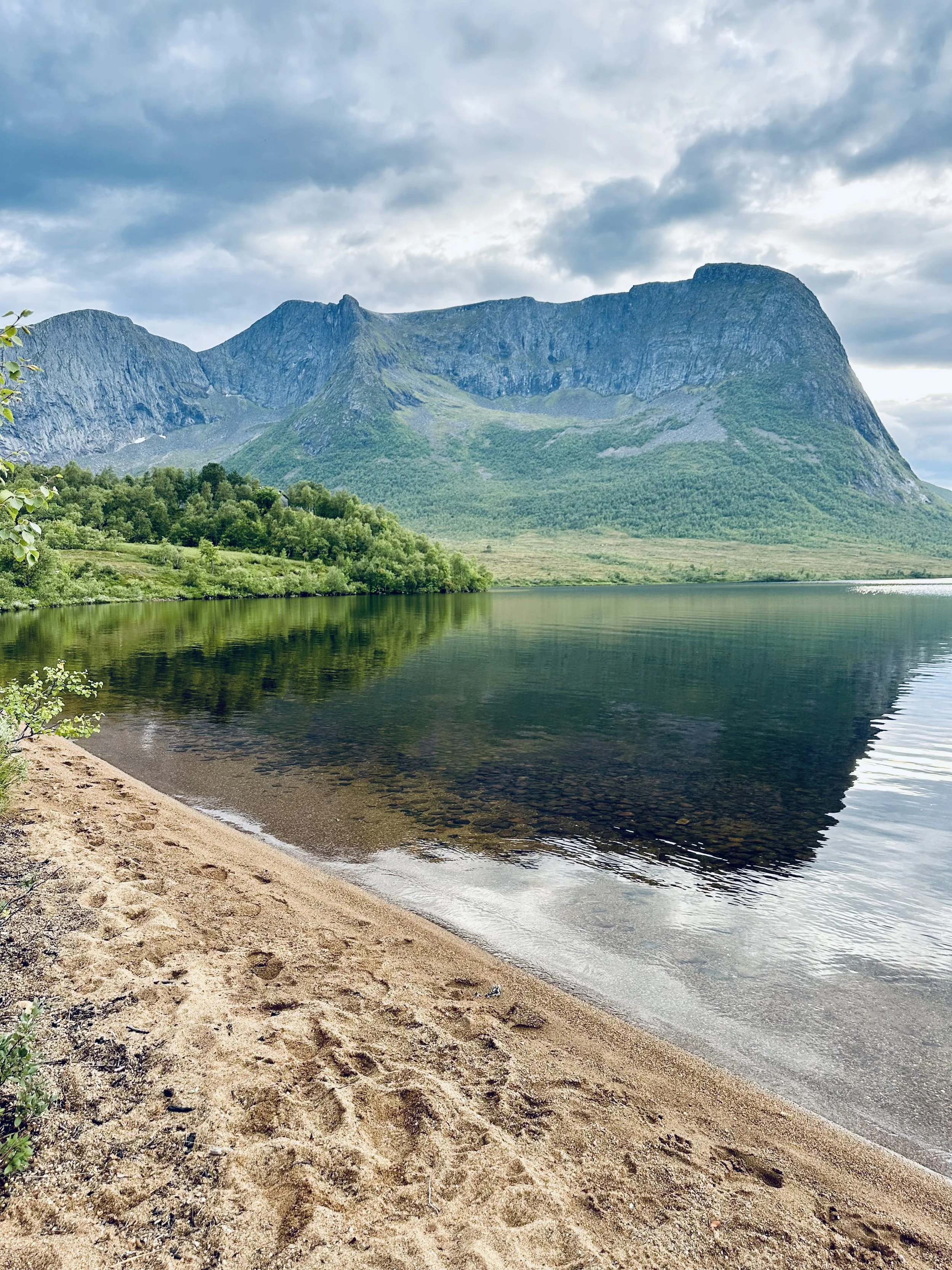 Sandstrand ved en innsjø med fjell i bakgrunnen under overskyet himmel