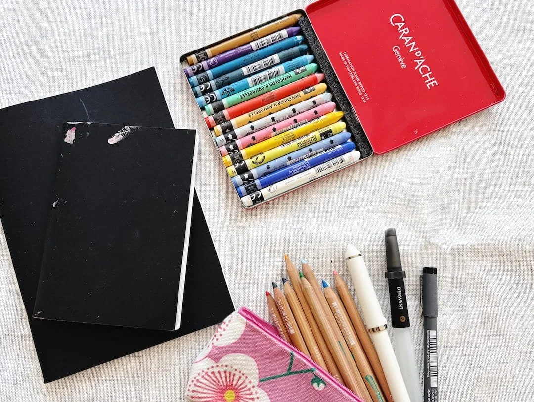 A small selection of art materials to make it easier to find time to create: image shows two black sketchbooks, a pencil case with pencils & pens and a small tin of colourful wax pastels on a table