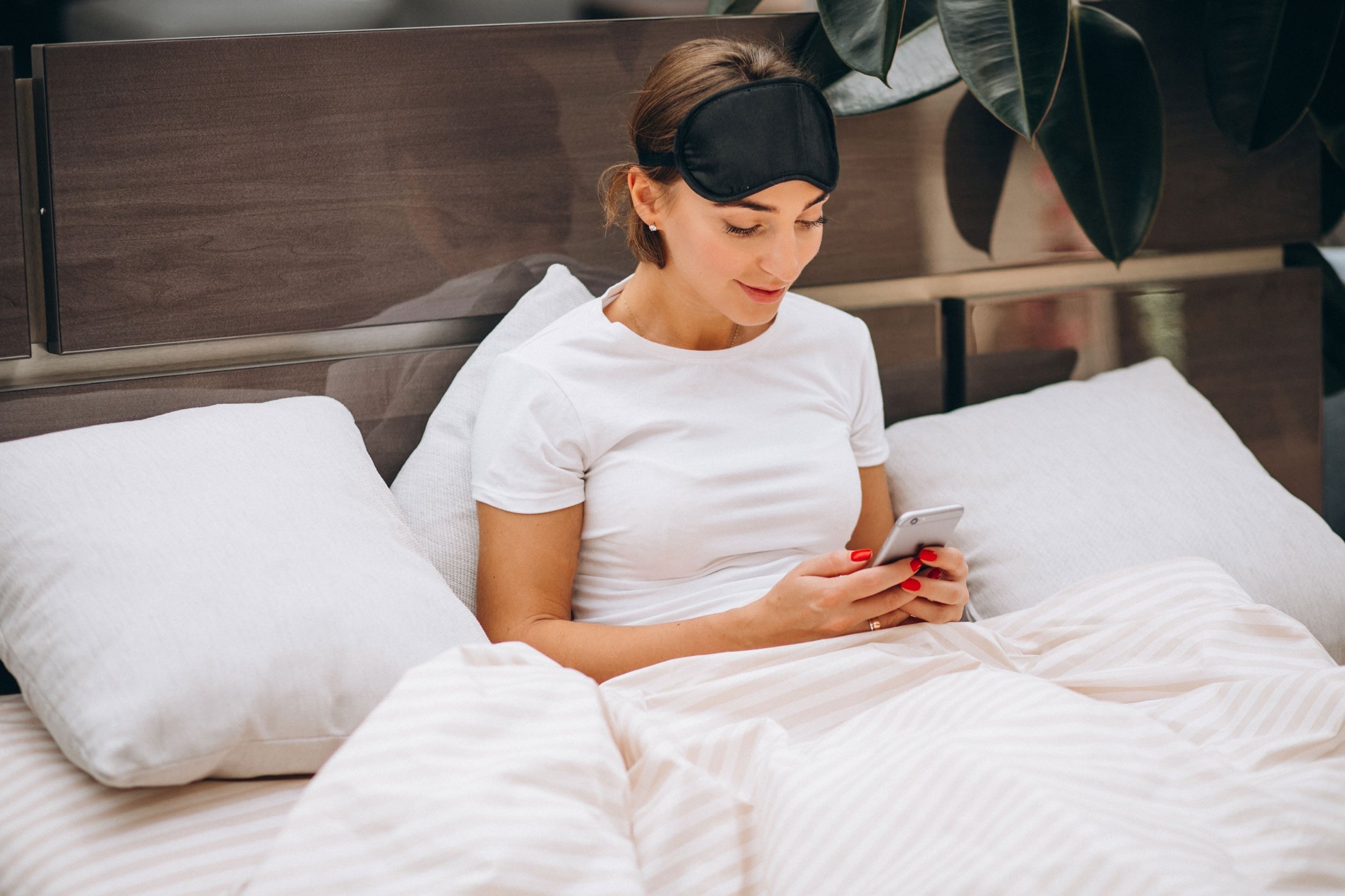  A woman with a sleep mask on her forehead, engrossed in her phone in bed. 