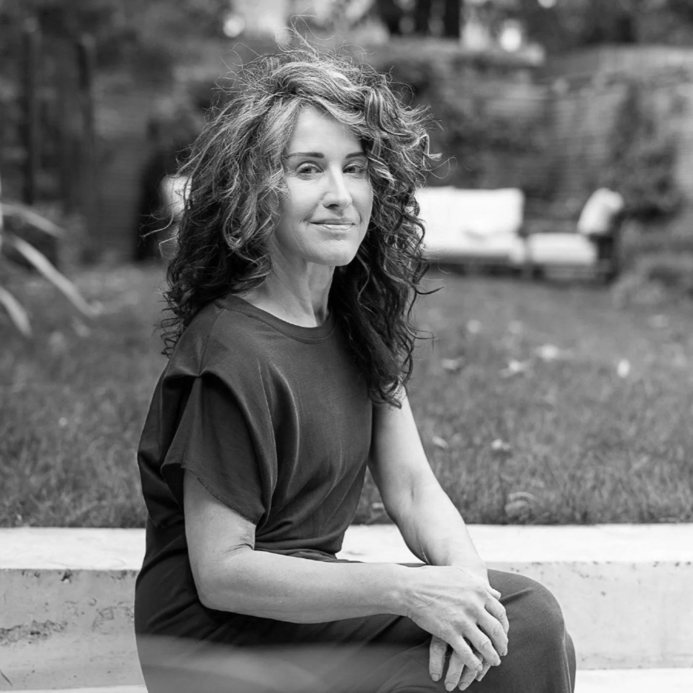 A black and white photo of a woman with curly hair sitting outdoors on a bench, with a blurred background of a park with grass and trees.