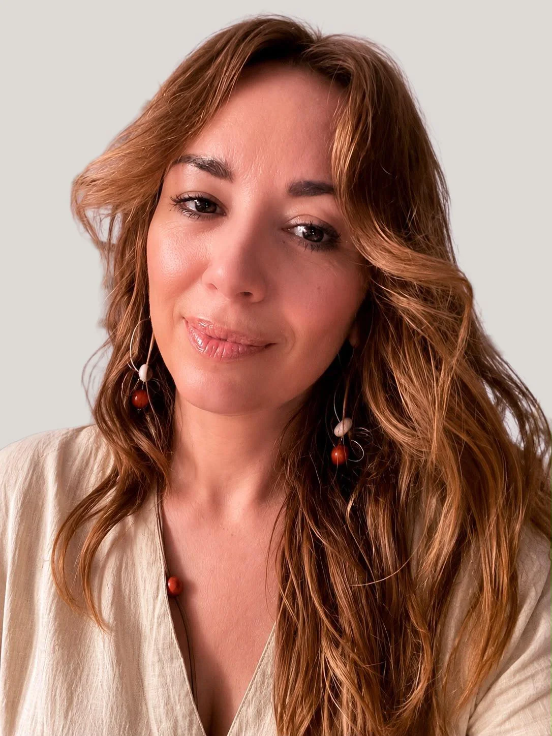 A woman with long, wavy auburn hair, wearing a neutral-colored top and unique earrings with beads and wire, smiling softly in front of a plain light background.