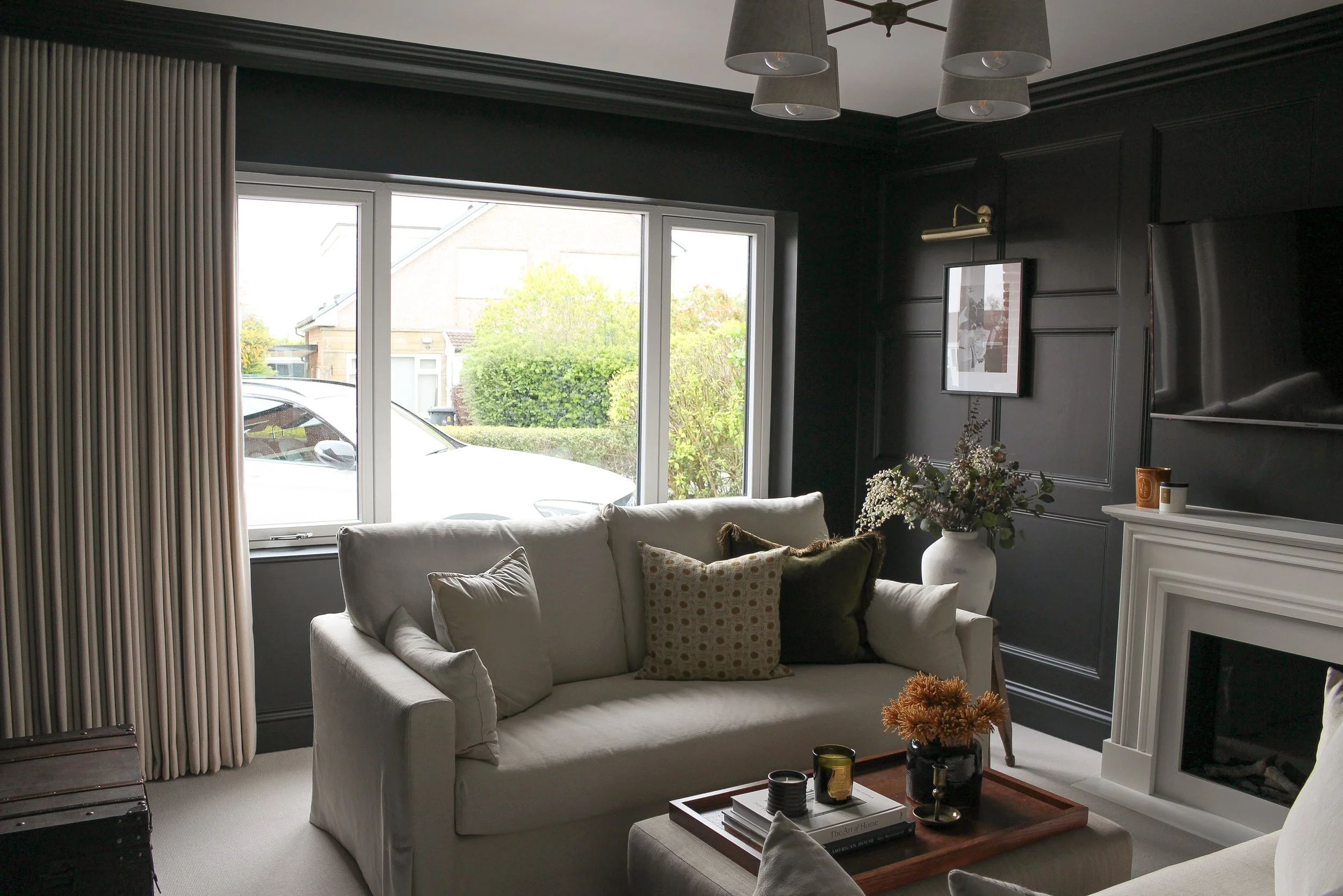 A cozy living room with a white sofa, throw pillows, a wooden tray on top of a ottoman coffee table with candles and flowers, black walls with framed art, a white fireplace mantel, a large window with beige curtains, and ceiling lights.