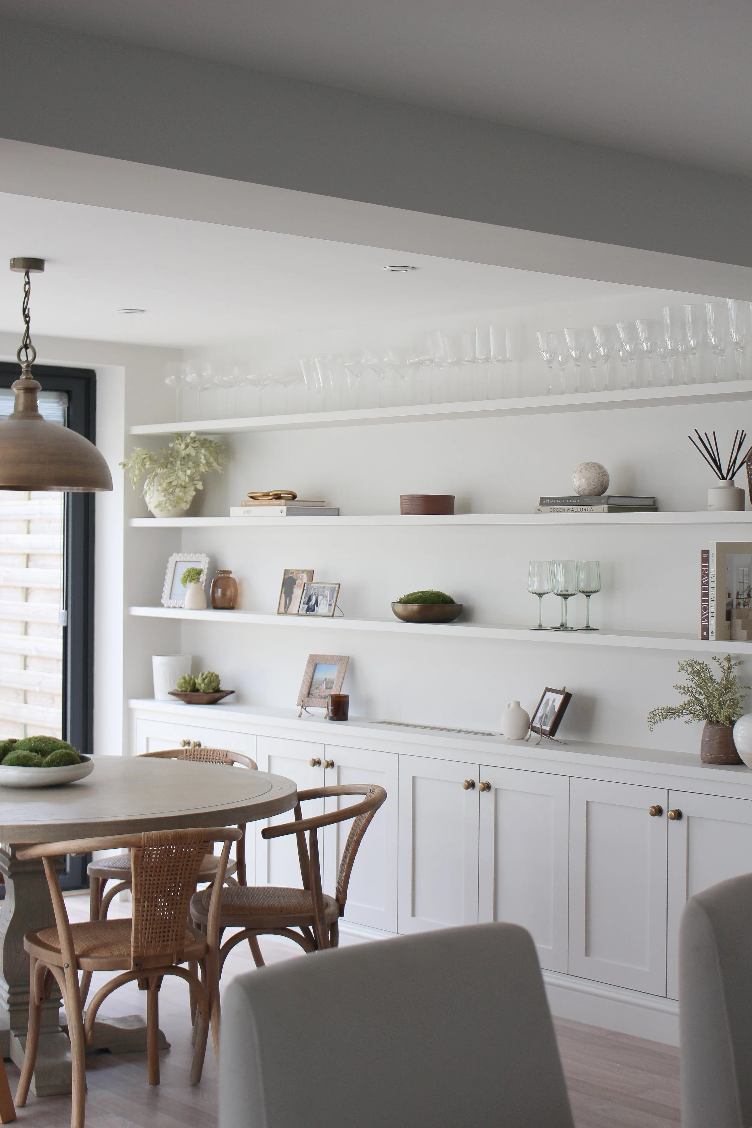 Modern dining room with white built-in cabinets and open shelves decorated with framed photos, books, vases, plants, and glassware, decorated in neutral tones.