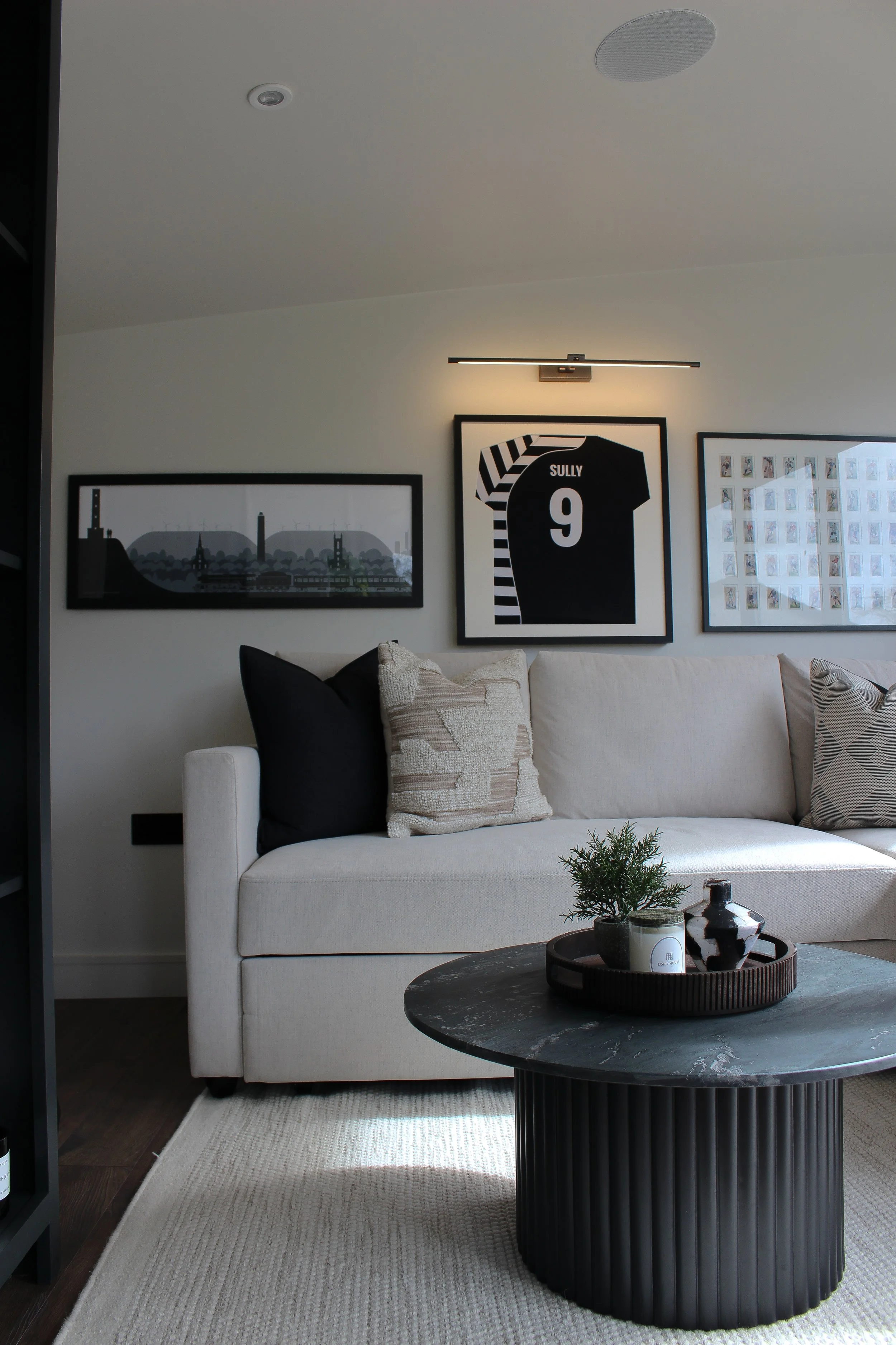 Office with a white sofa, decorative pillows, a black round coffee table with a plant and decorative items, and artwork on the wall including a framed rugby top. Garden office designed by Yazzmin Lovelle Interiors.