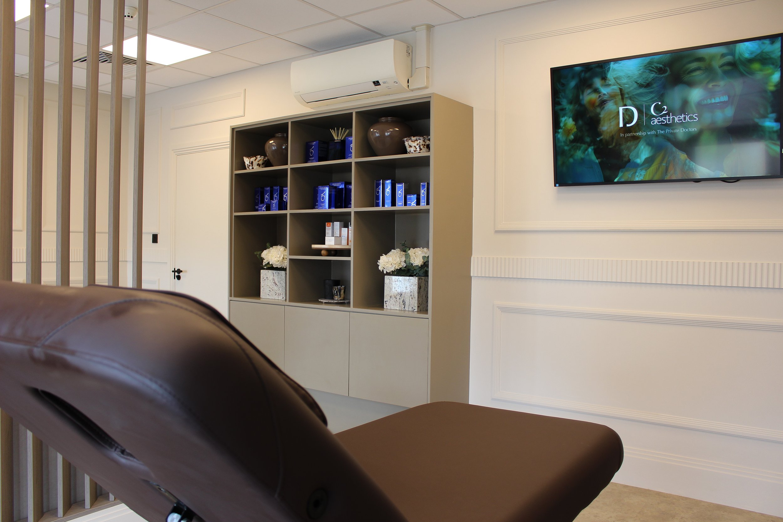 Interior of a aesthetics clinic with a brown treatment chair, a shelving unit with zo skincare and decorative items, a flat-screen TV on the wall displaying the logo c2 Aesthetics,' and a white air conditioning unit.