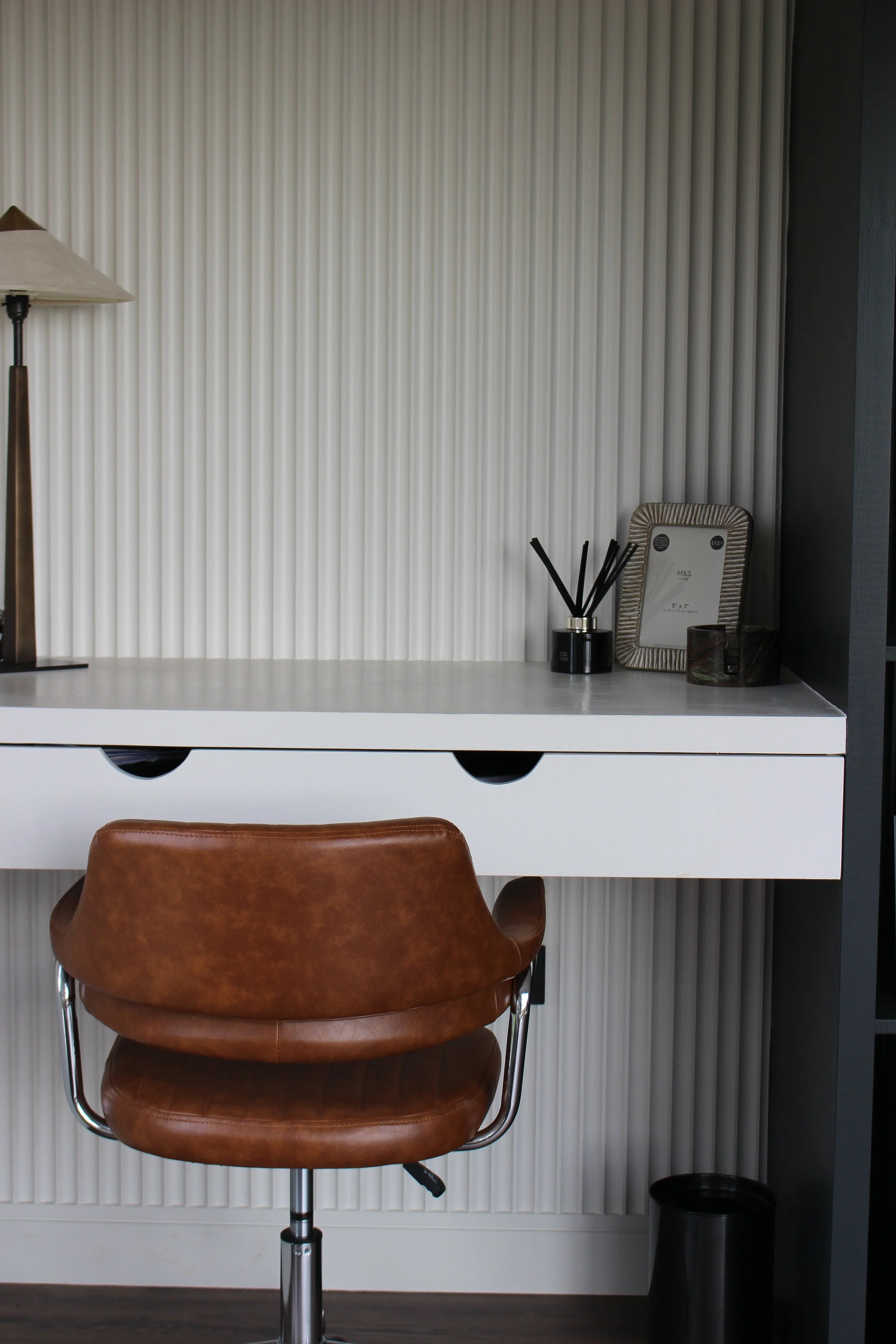 A modern workspace with a brown leather office chair in front of a white desk, a picture frame, and reed diffusers on the desk, against a textured white wall. Garden office designed by Yazzmin Lovelle Interiors.