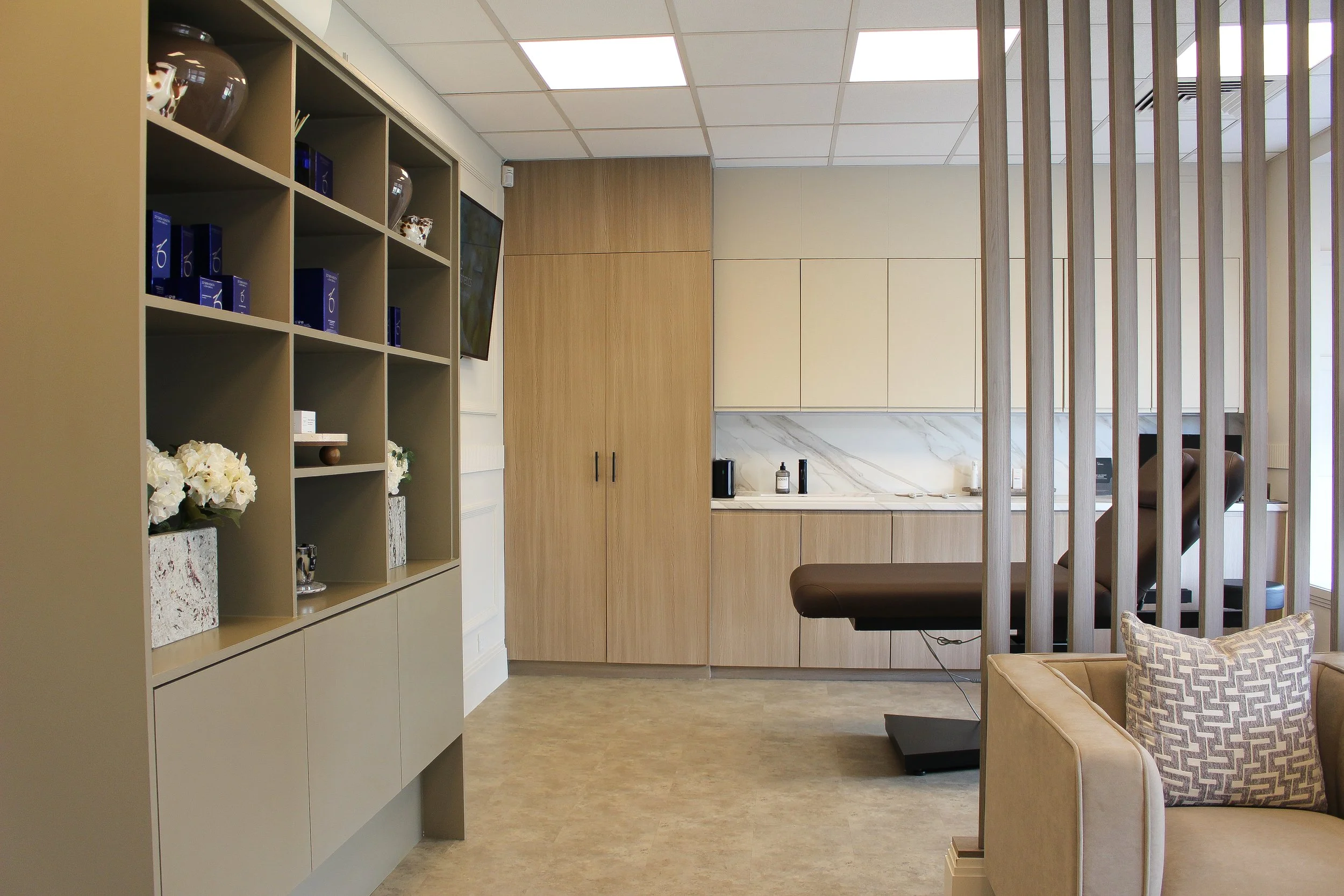 A medical or aesthetic consultation room with medical examination table, wooden cabinetry, decorative shelves with flowers, and a seating area with a patterned pillow. Clinic designed by Yazzmin Lovelle Interiors