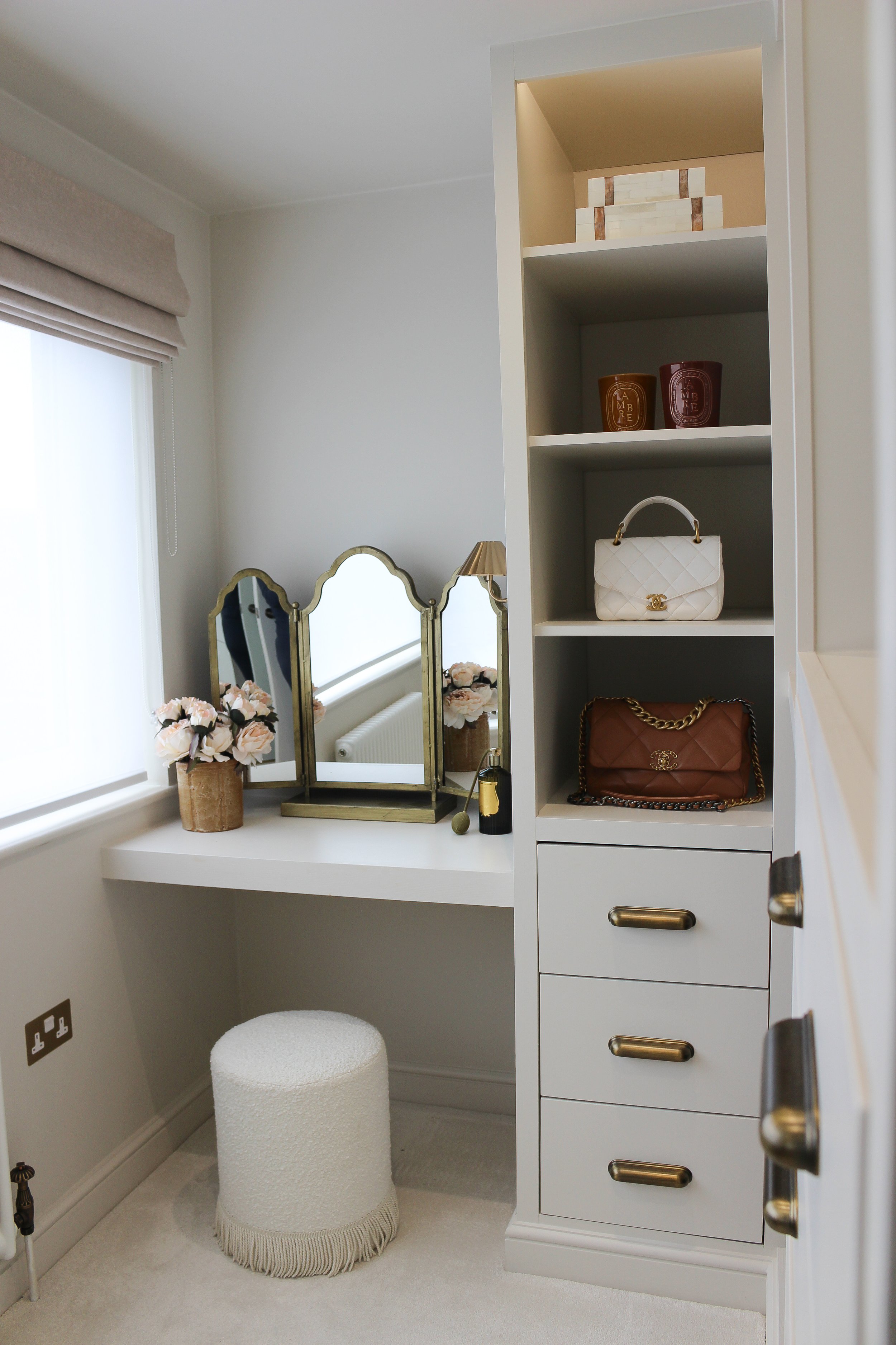 Luxury vanity area. Antique brass mirror sits on top of the dressing table, with a stone vase with pink flowers. there is also a brass wall light and dressing room shelving with built in draws displaying designer handbags 