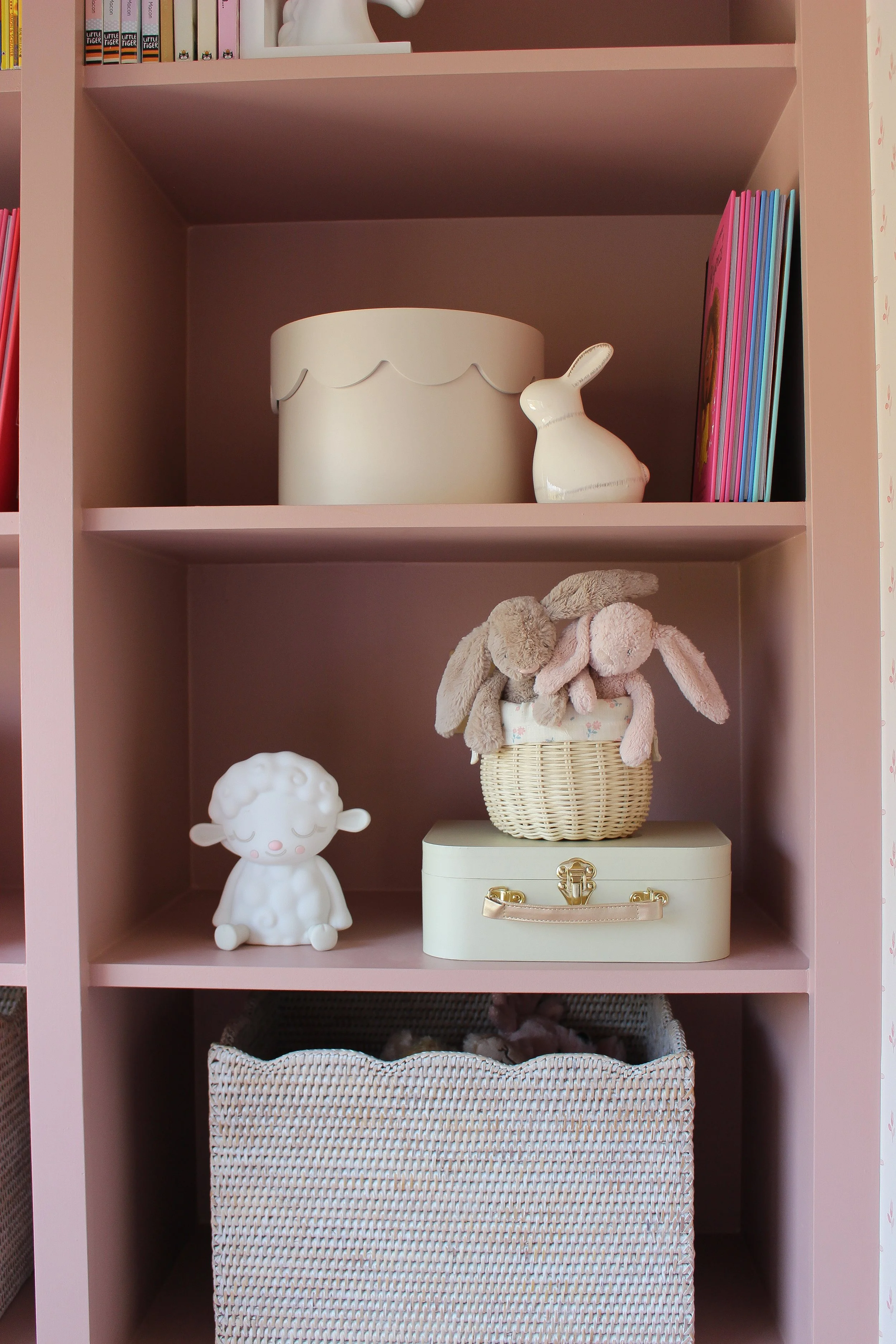 Pink shelf with toys, books, and storage boxes, including a bunny figurine, stuffed animals, and a white decorative box. Girls bedroom designed by Yazzmin Lovelle Interiors