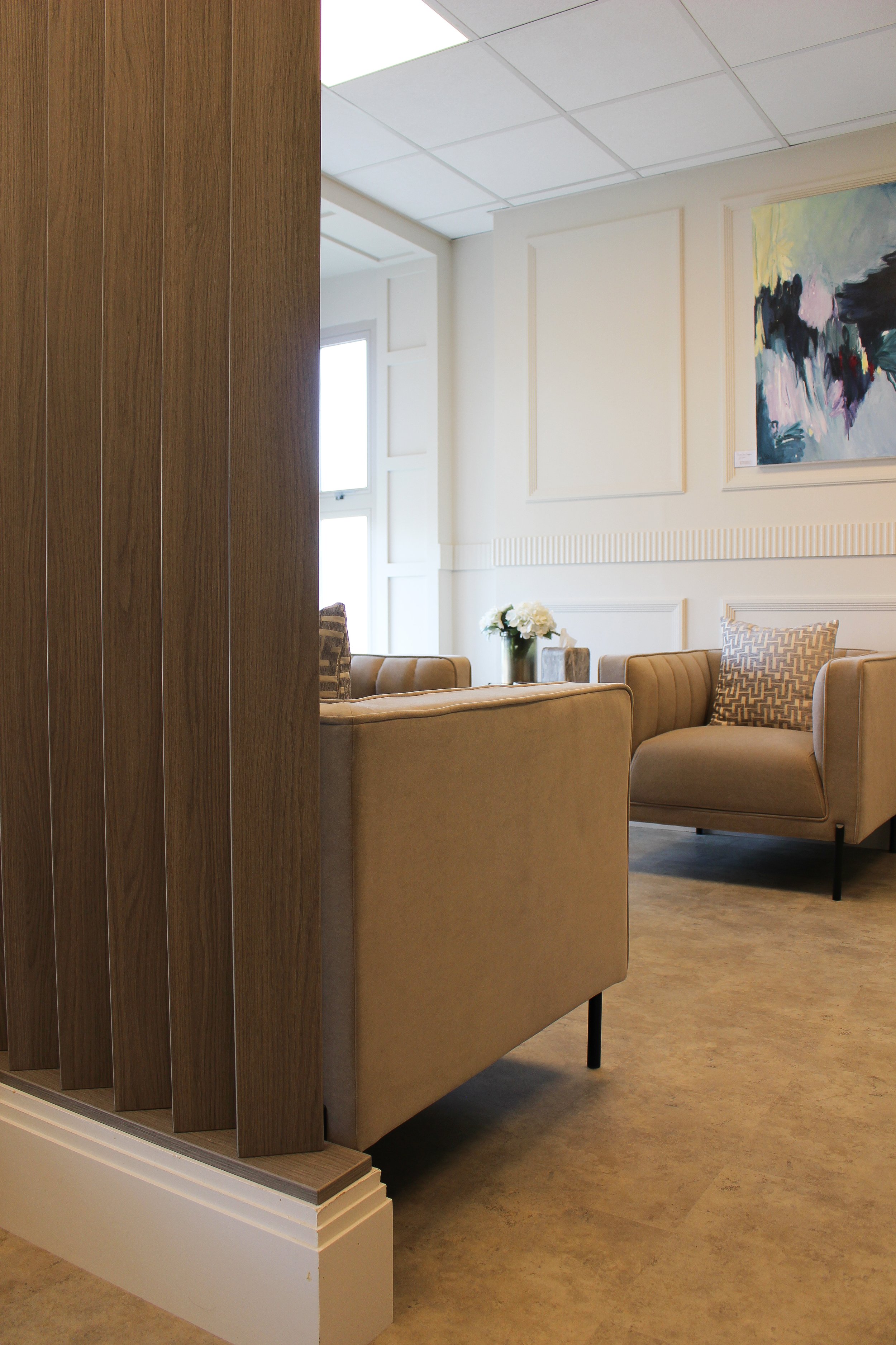 A luxury waiting area with beige armchairs, decorative pillows, a vase with white flowers, abstract artwork on the wall, large windows letting in natural light, and a wooden partition. Clinic designed by Yazzmin Lovelle Interiors