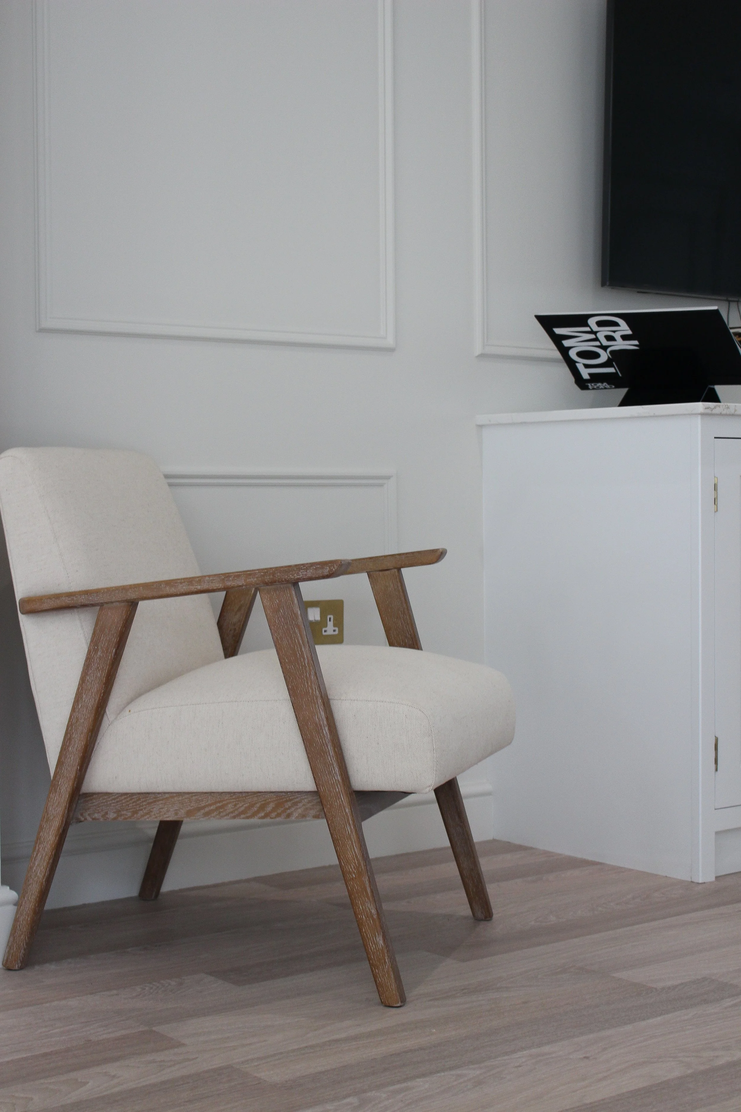 A beige upholstered armchair with wooden armrests and legs sitting on a light wood floor in a room with white walls, a white cabinet, and a wall-mounted TV.