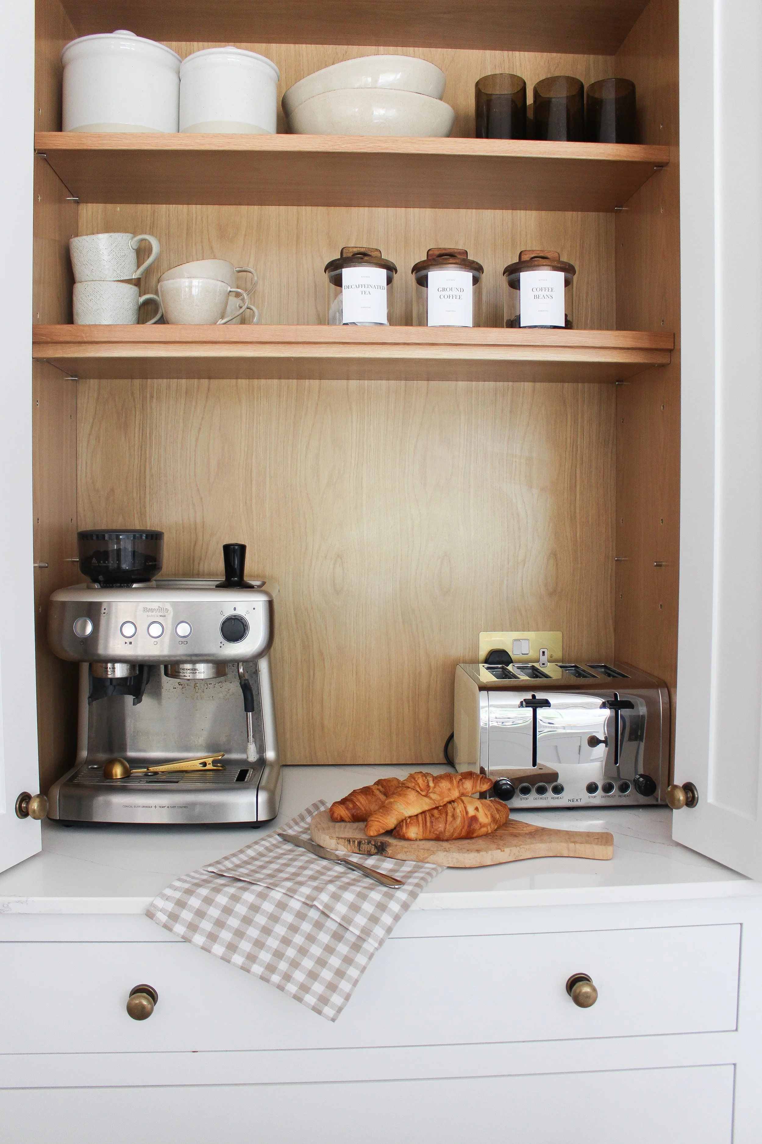 A breakfast setup with croissants on a wooden cutting board, a beige and white checkered napkin, a stovetop espresso machine, and a toaster in a kitchen cabinet with shelves holding bowls, cups, and jars.