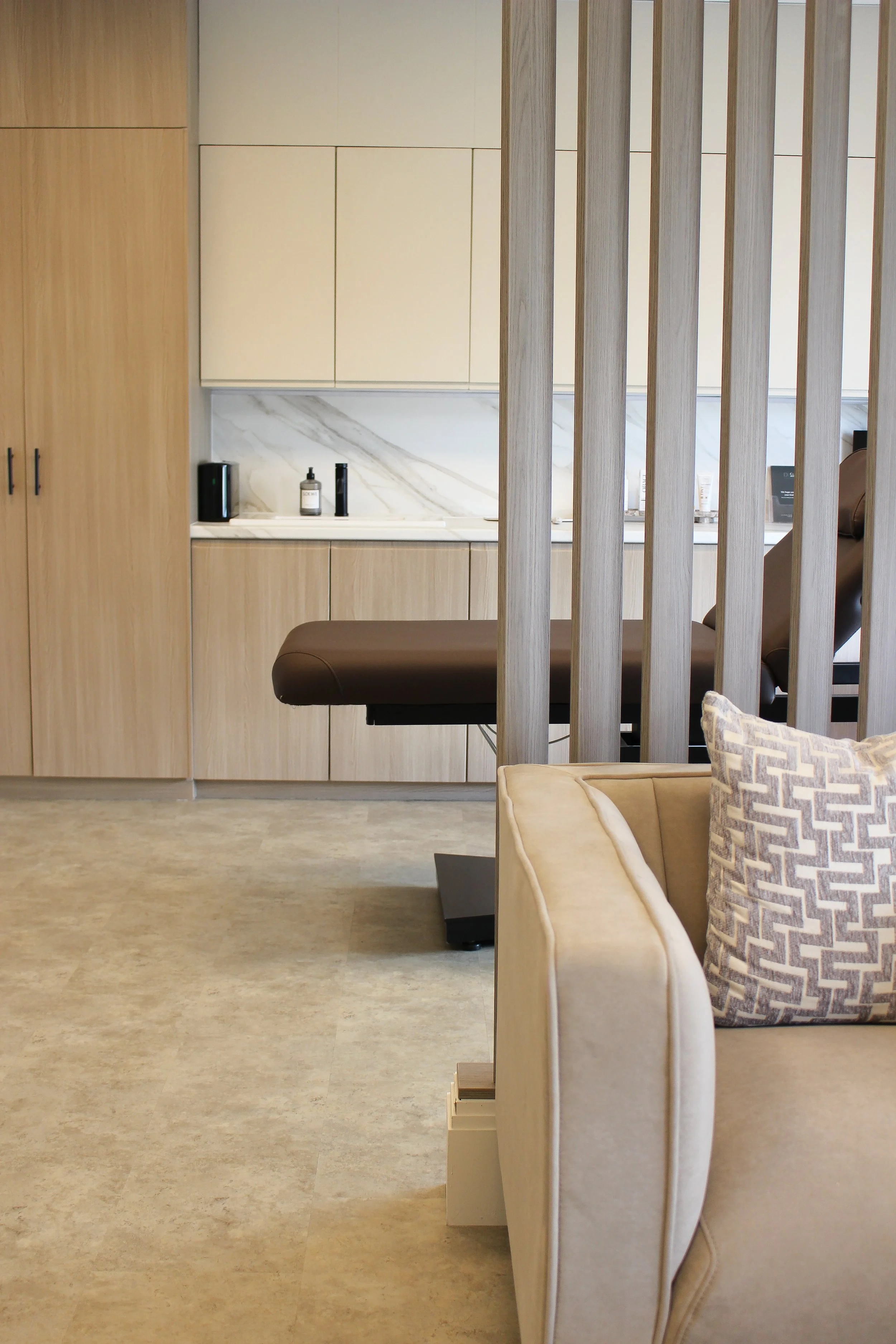 Clinic Waiting area with beige chair, patterned pillow, wooden slatted divider, and a treatment room with treatment table in the background. Clinic designed by Yazzmin Lovelle Interiors 