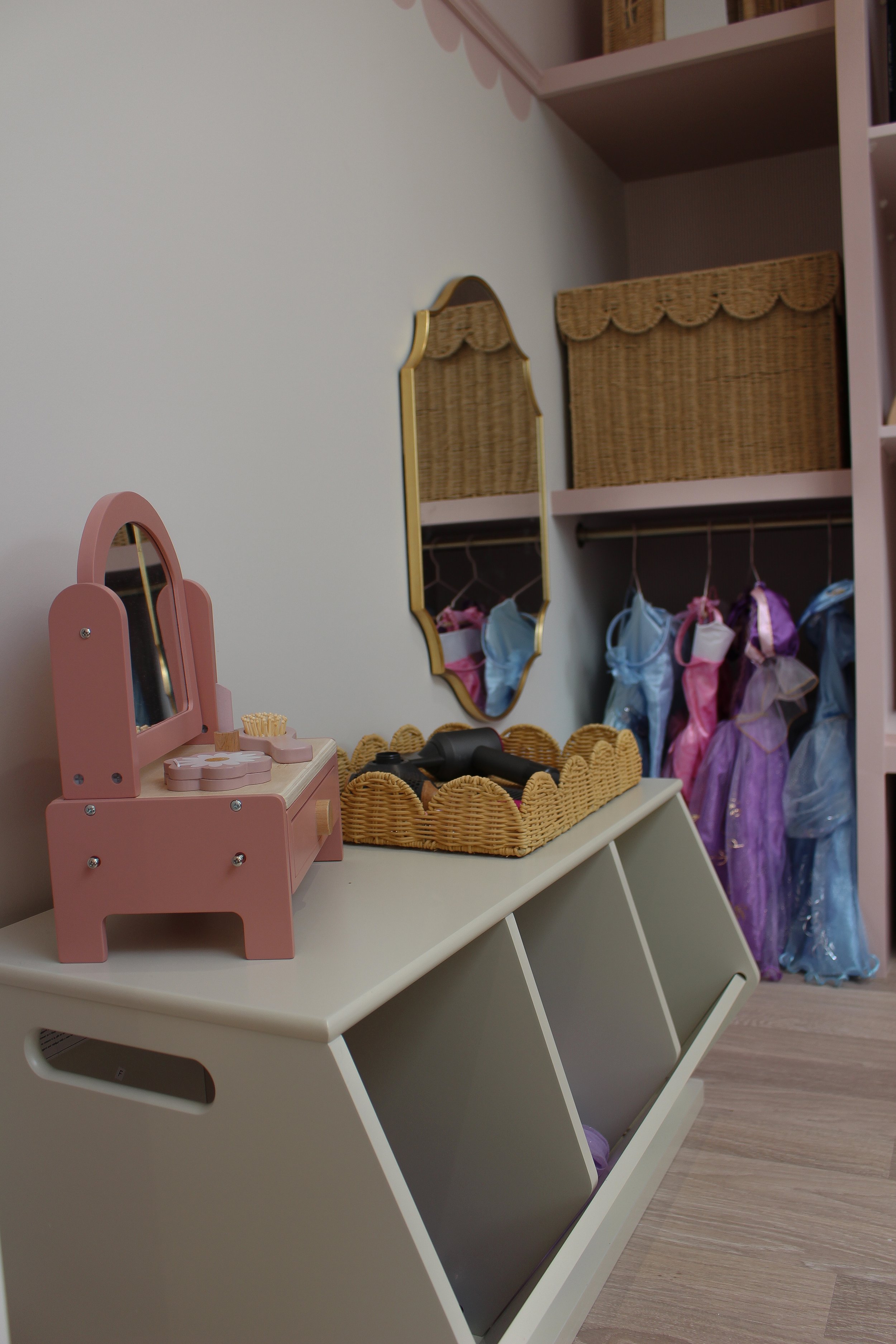 A children's dressing area with a white storage bench, pink toy vanity, wicker tray with black hair dryer, and a wall with hanging princess dresses in purple, pink, and blue. Playroom designed by Yazzmin Lovelle Interiors.