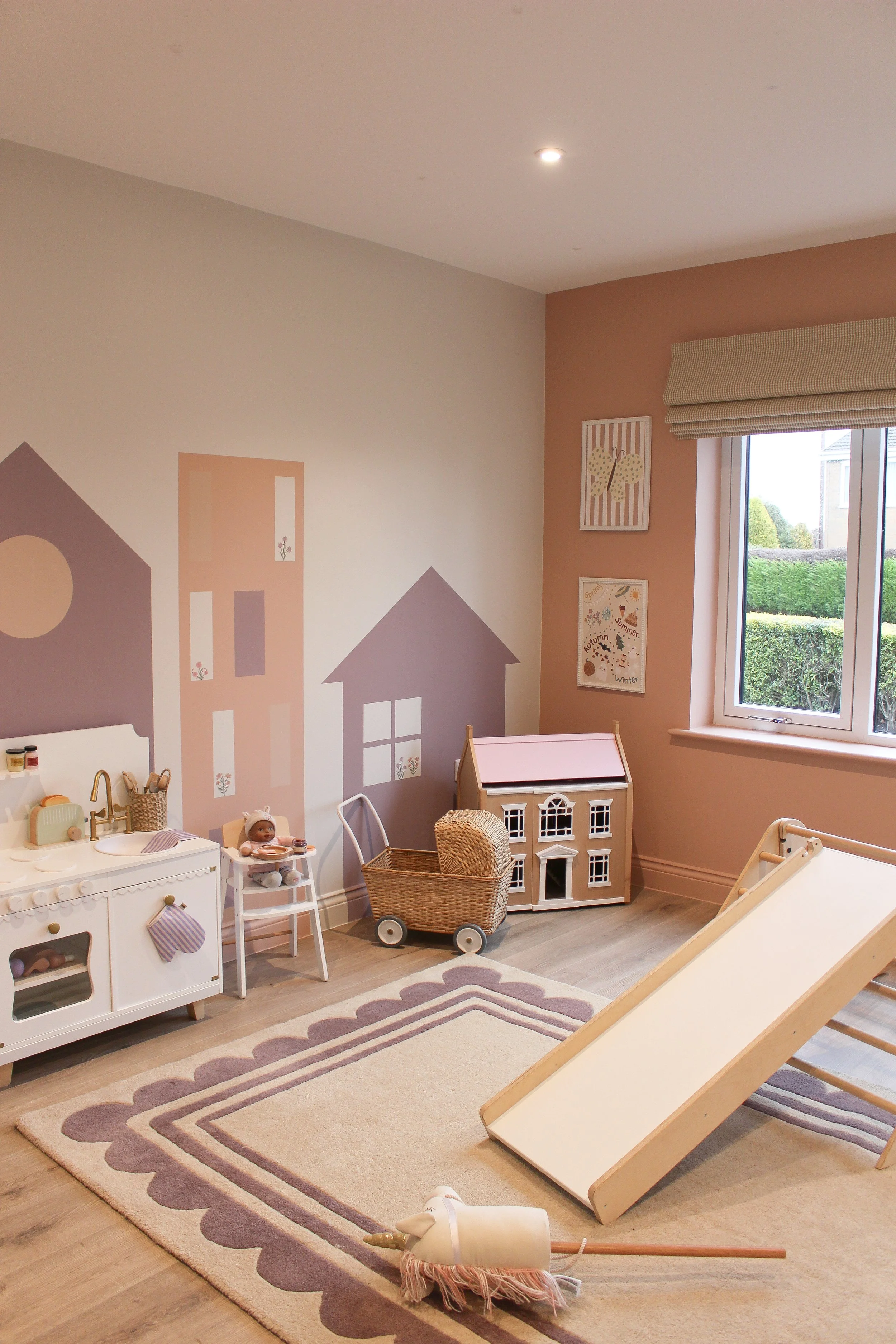 Decorated children's playroom or nursery featuring pastel coloured walls, wall art, a mini kitchen set, a doll, a toy house, a wagon, a slide, and a plush unicorn toy on the floor. Playroom designed by Yazzmin Lovelle Interiors