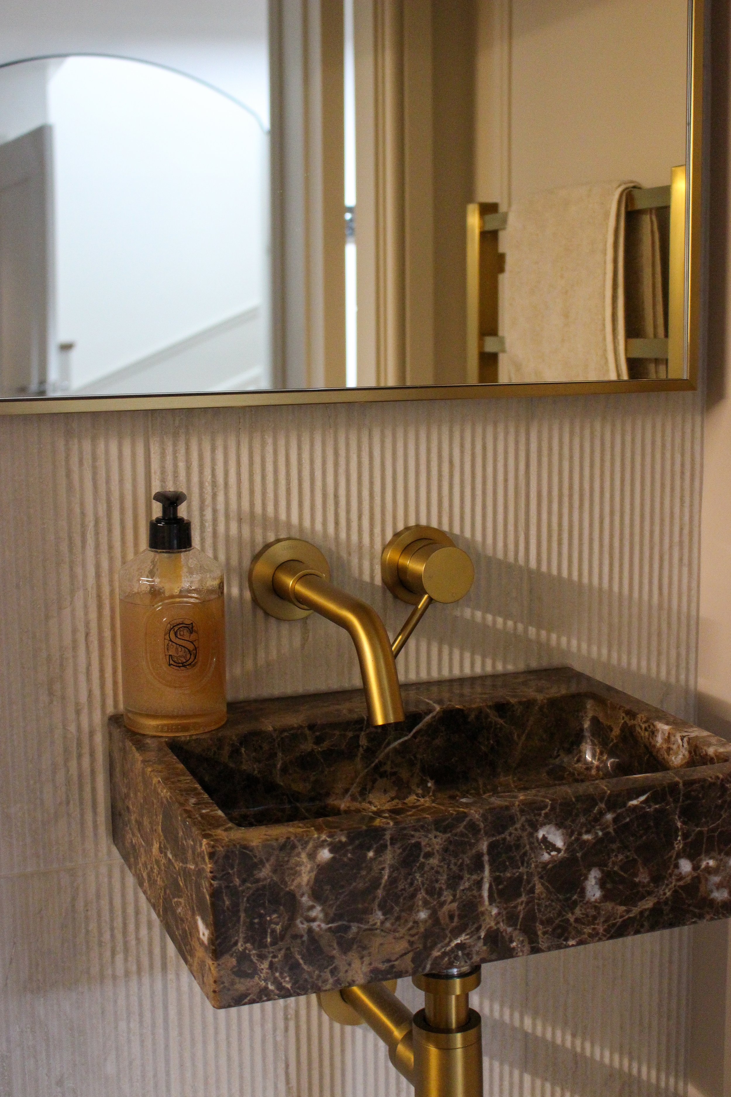 Small dark marble bathroom sink with gold tap and soap dispenser, mirror above, textured tile wall behind, with a reflection of doorway and staircase in the mirror. Downstairs toilet designed by Yazzmin Lovelle Interiors 