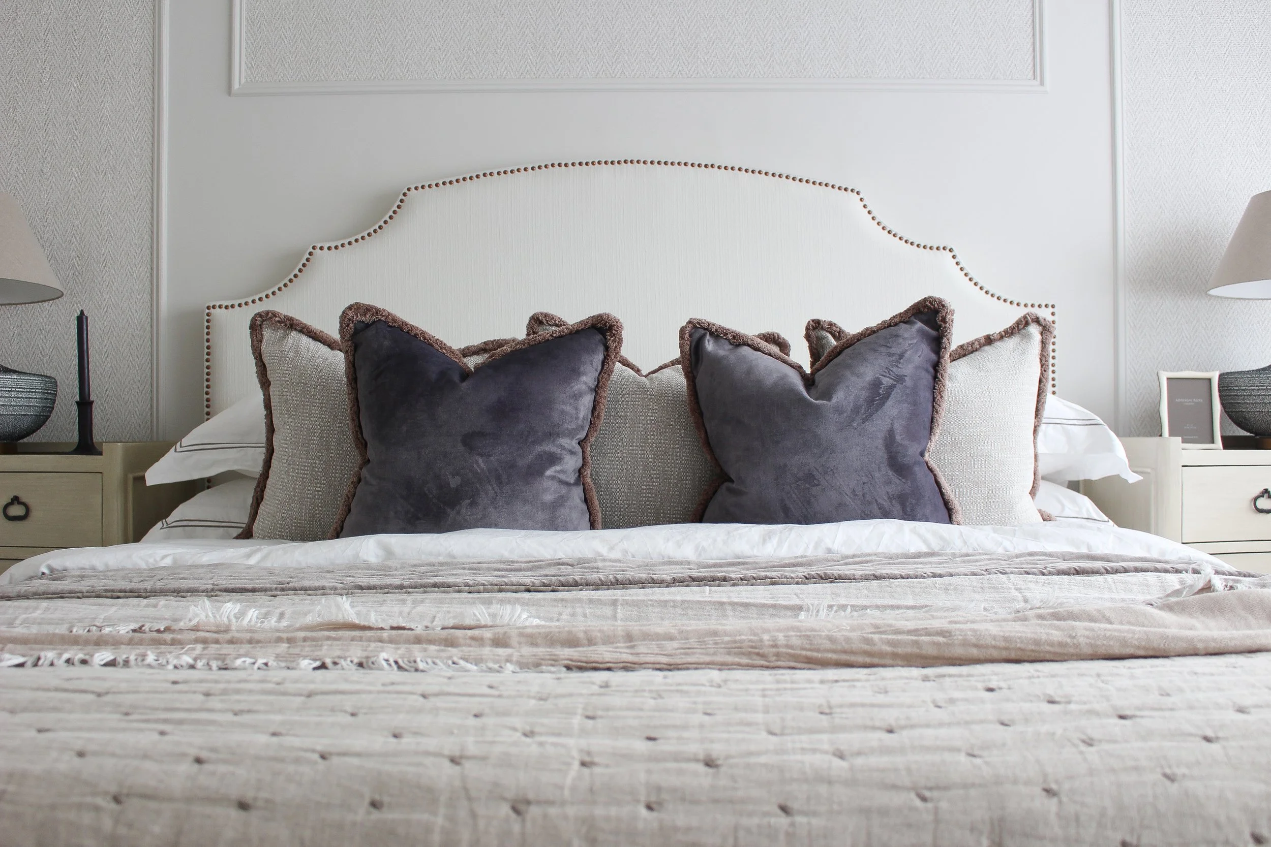 A neatly made bed with a cream coloured headboard and luxury throw cushions, including three large dark grey velvet pillows with brown trim, in a bedroom with textured panelled walls, large bedside tables with lamps, and a small photo frame.