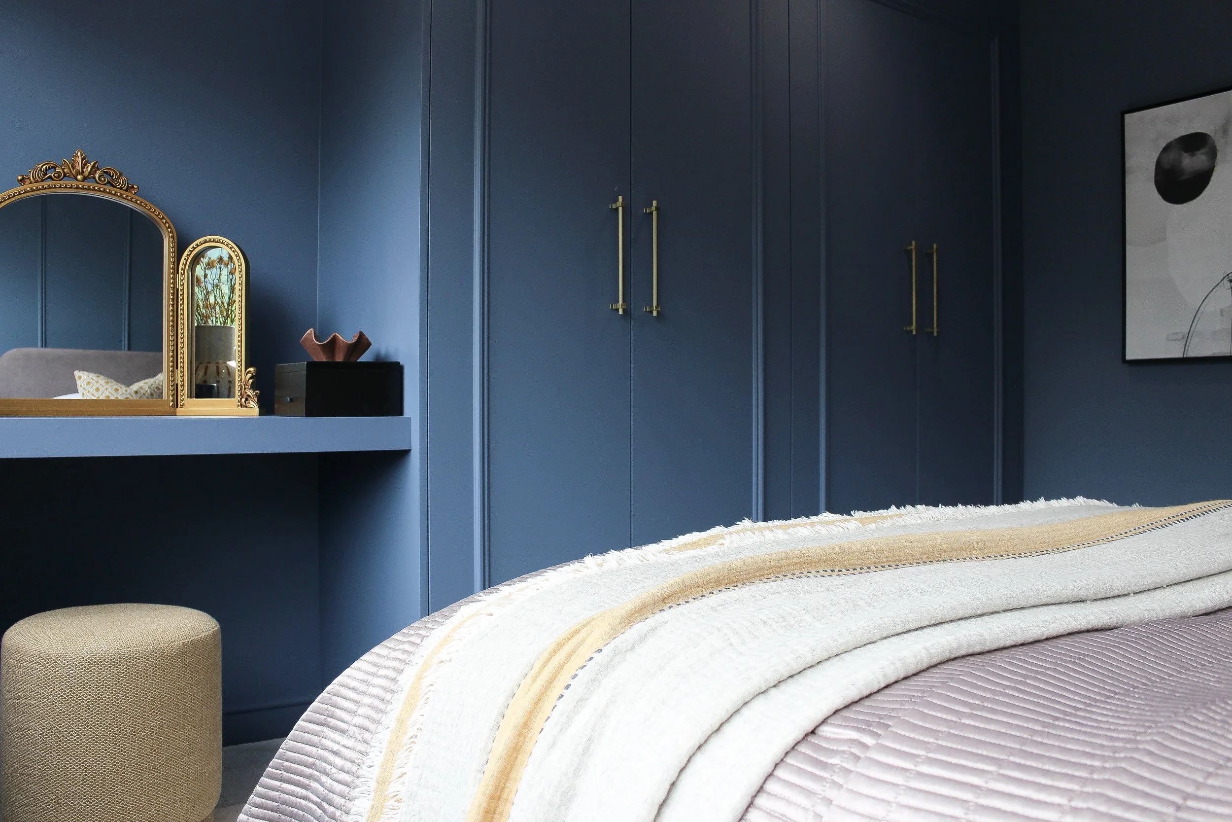 A bedroom with blue walls, a bed covered with beige and cream bedding, a beige ottoman, and blue cabinets with gold handles. A mirror with a gold frame, a small black table with a decorative bowl, and a large abstract black and white painting are als