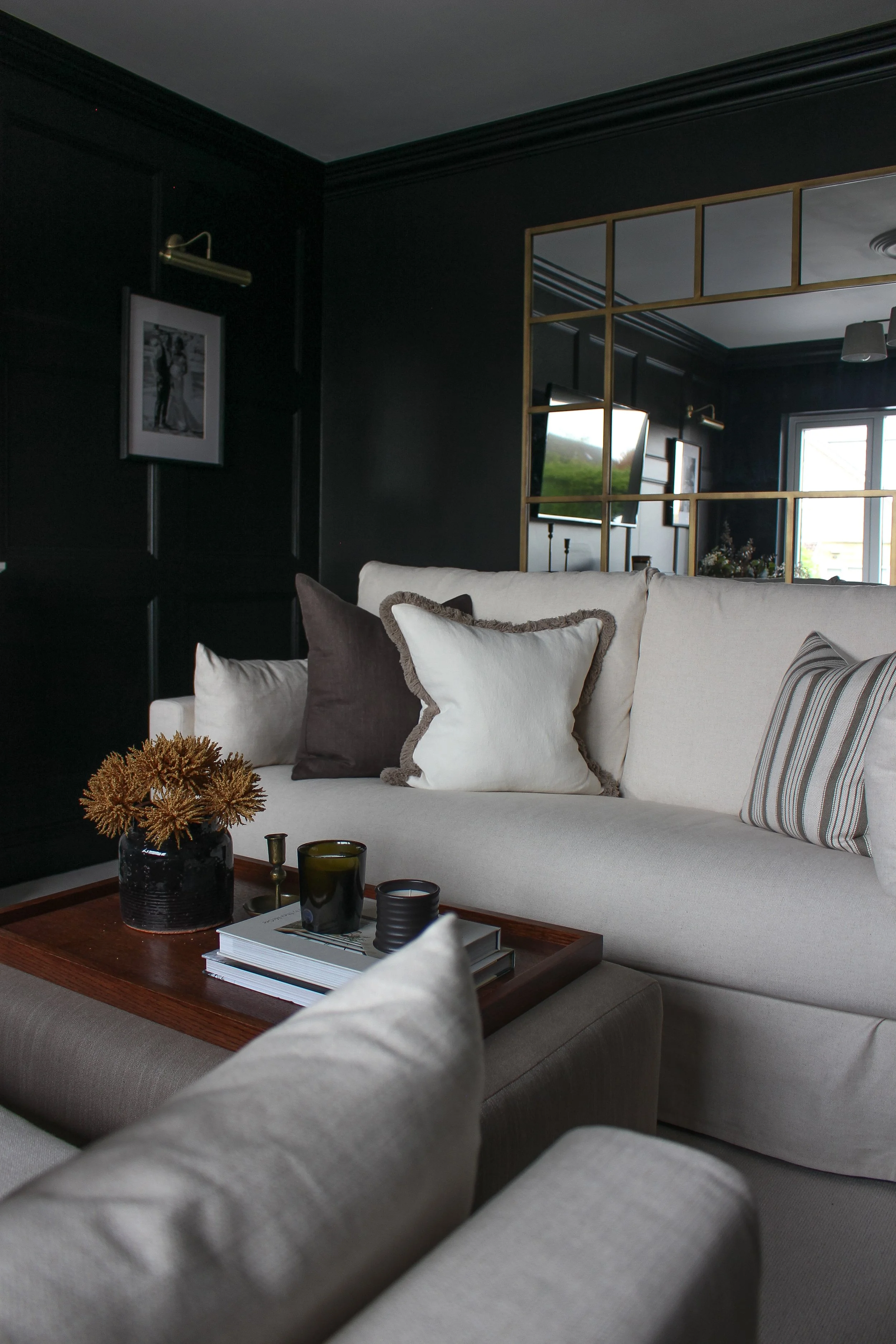 A living room with a white sofa, decorative pillows, a wooden tray on top of a fabric coffee table with candles and a vase, and dark panelled walls with framed black-and-white photos. Living room designed by Yazzmin Lovelle Interiors.