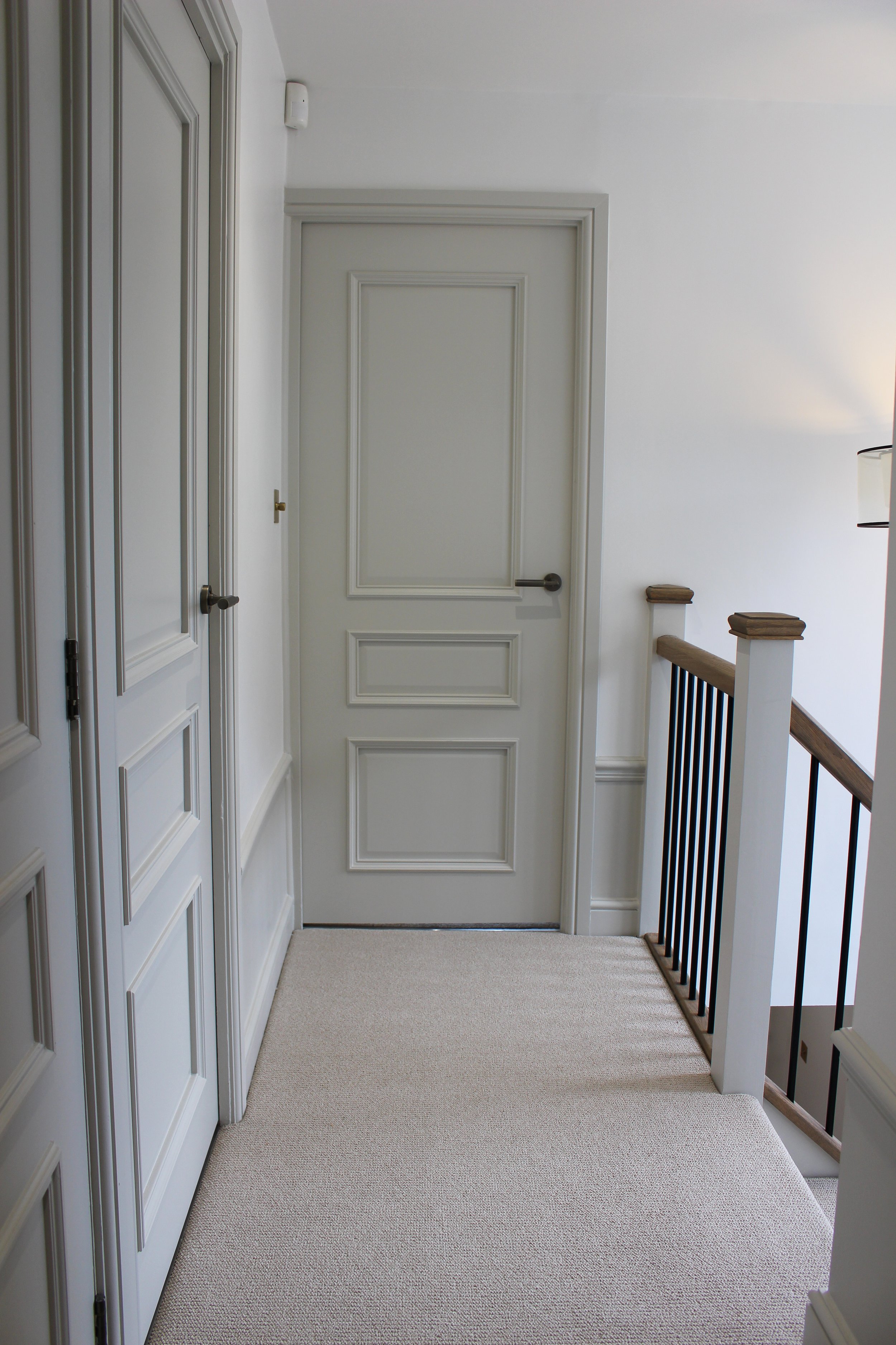 An upstairs hallway with beige textured carpet, white walls, a white door with decorative moulding, a staircase with a wooden handrail and black metal balusters, and ceiling pendant over the stairs. Hallway designed by Yazzmin Lovelle Interiors. 