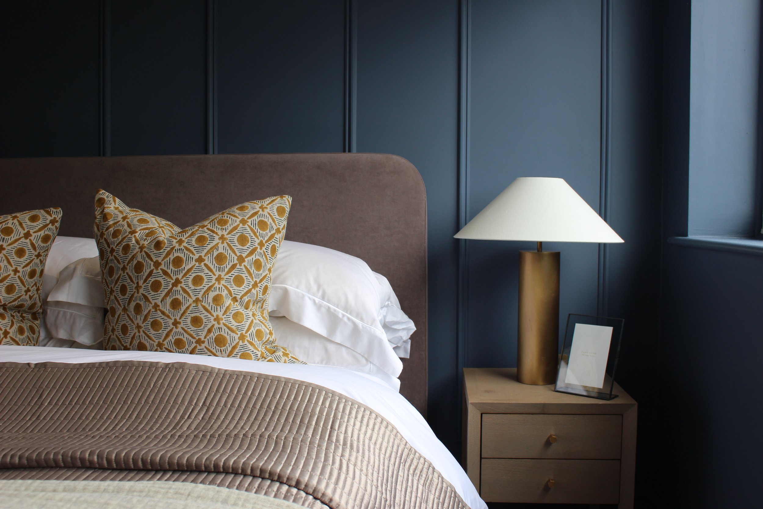 A bedroom with a grey upholstered headboard, patterned yellow and grey throw cushions, a beige quilted blanket, a wooden bedside table with a gold lamp, and a framed picture, against dark blue panelled walls.
