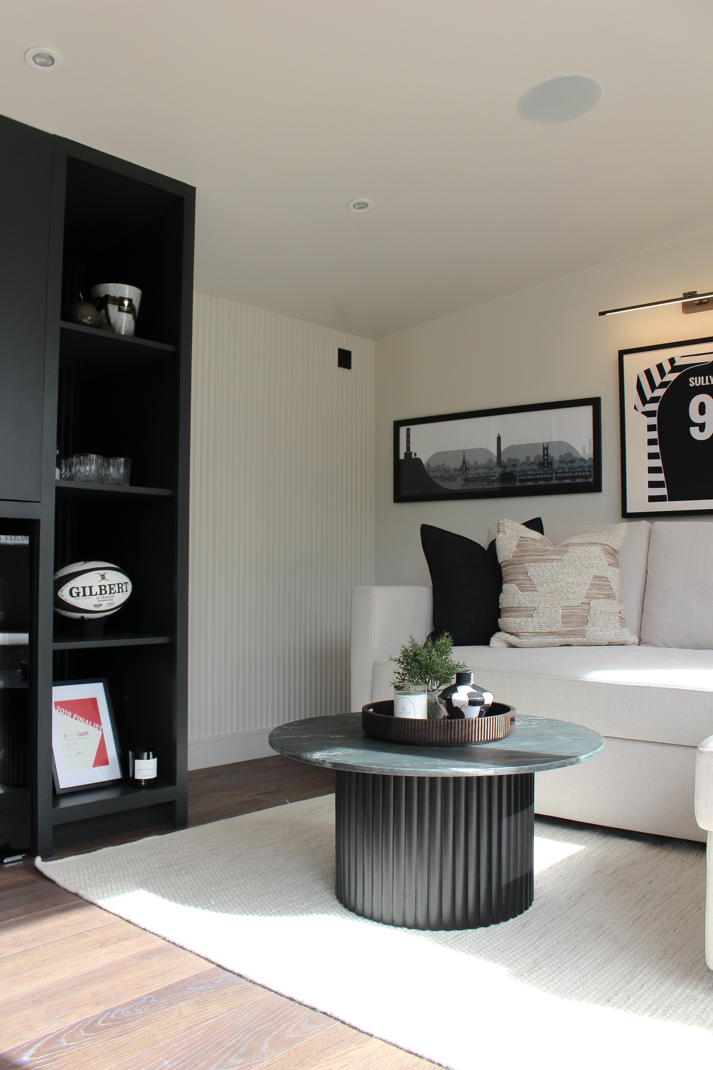 Office with a black circular coffee table, white sofa with black and beige cushions, wall art, and a black shelving unit with decorative items and a Gilbert rugby ball. Garden office designed by Yazzmin Lovelle Interiors.