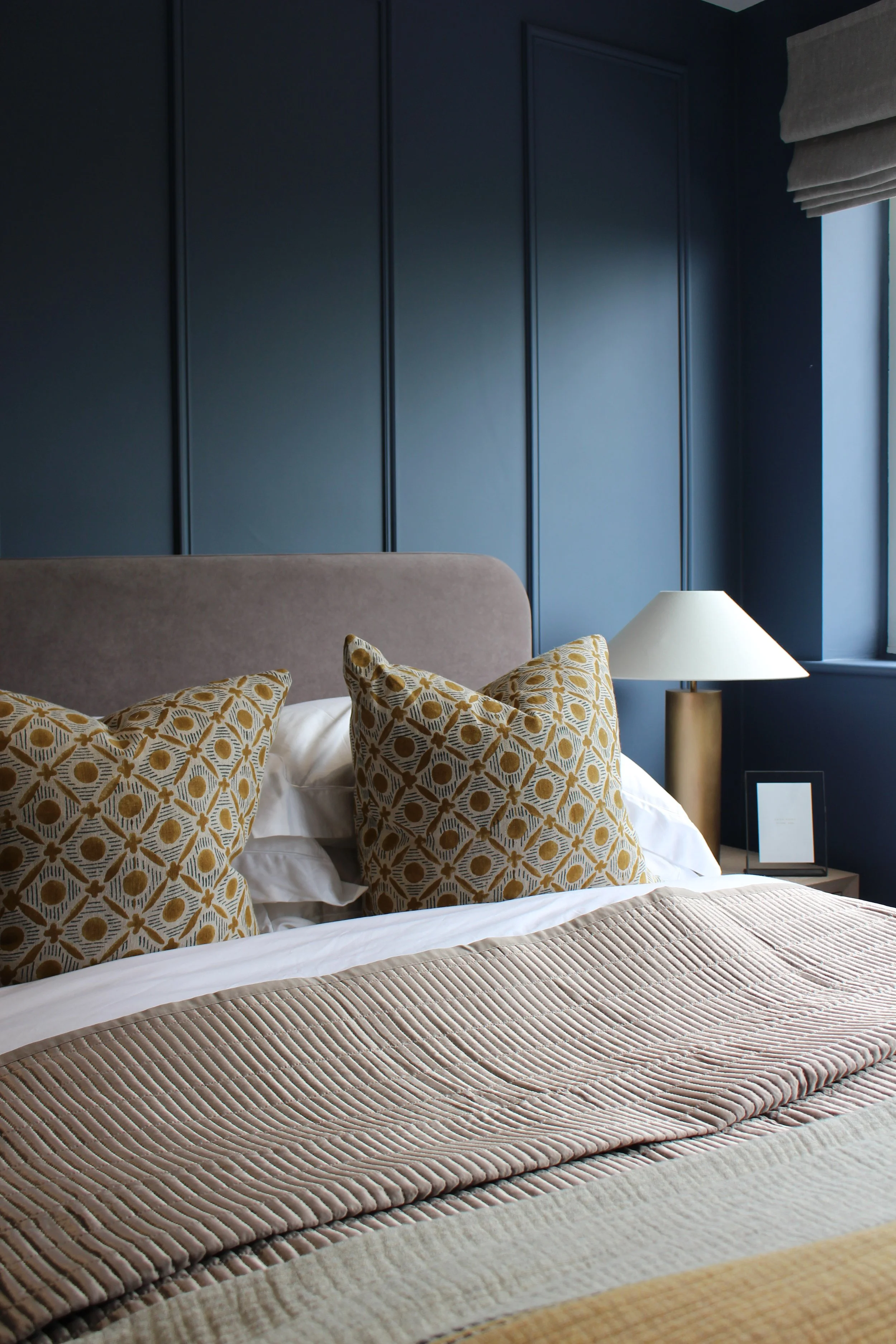 A neatly made bed with beige and patterned throw pillows in a bedroom with dark blue wall paneling, a white lamp on the side table, and a window with linen blinds. Guest Bedroom designed by Yazzmin Lovelle Interiors.