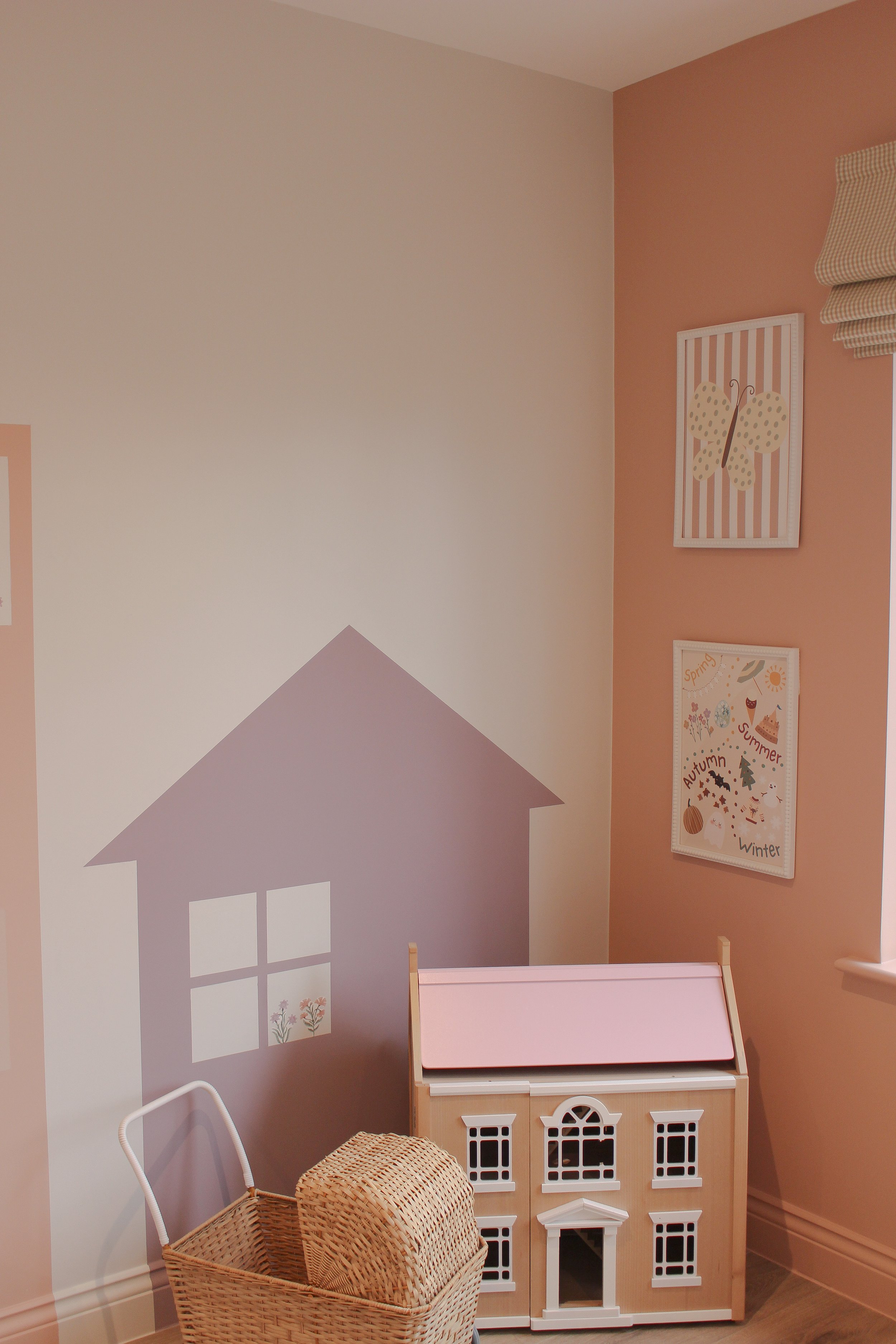 A corner of a child's playroom with pink and beige walls. There is a dollhouse with a pink roof, a wicker basket, and wall art that features a butterfly and a seasonal chart with the words 'Spring,' 'Summer,' 'Autumn,' and 'Winter'.