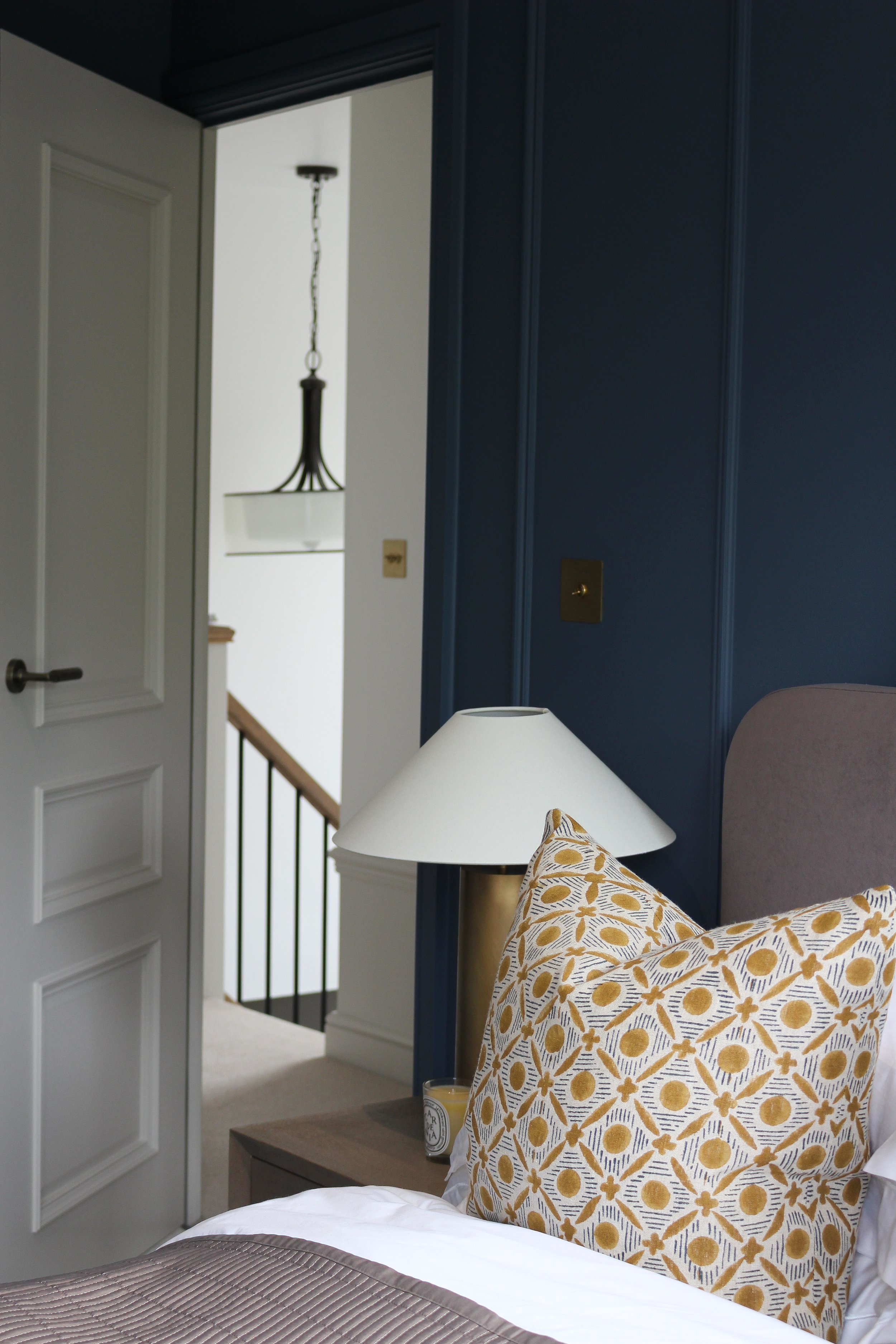 A bedroom with a wooden bedside table with a gold lamp and cushion on the bed, an open door, a staircase landing with a hanging light fixture, and a blue panelled wall. Guest bedroom designed by Yazzmin Lovelle Interiors 