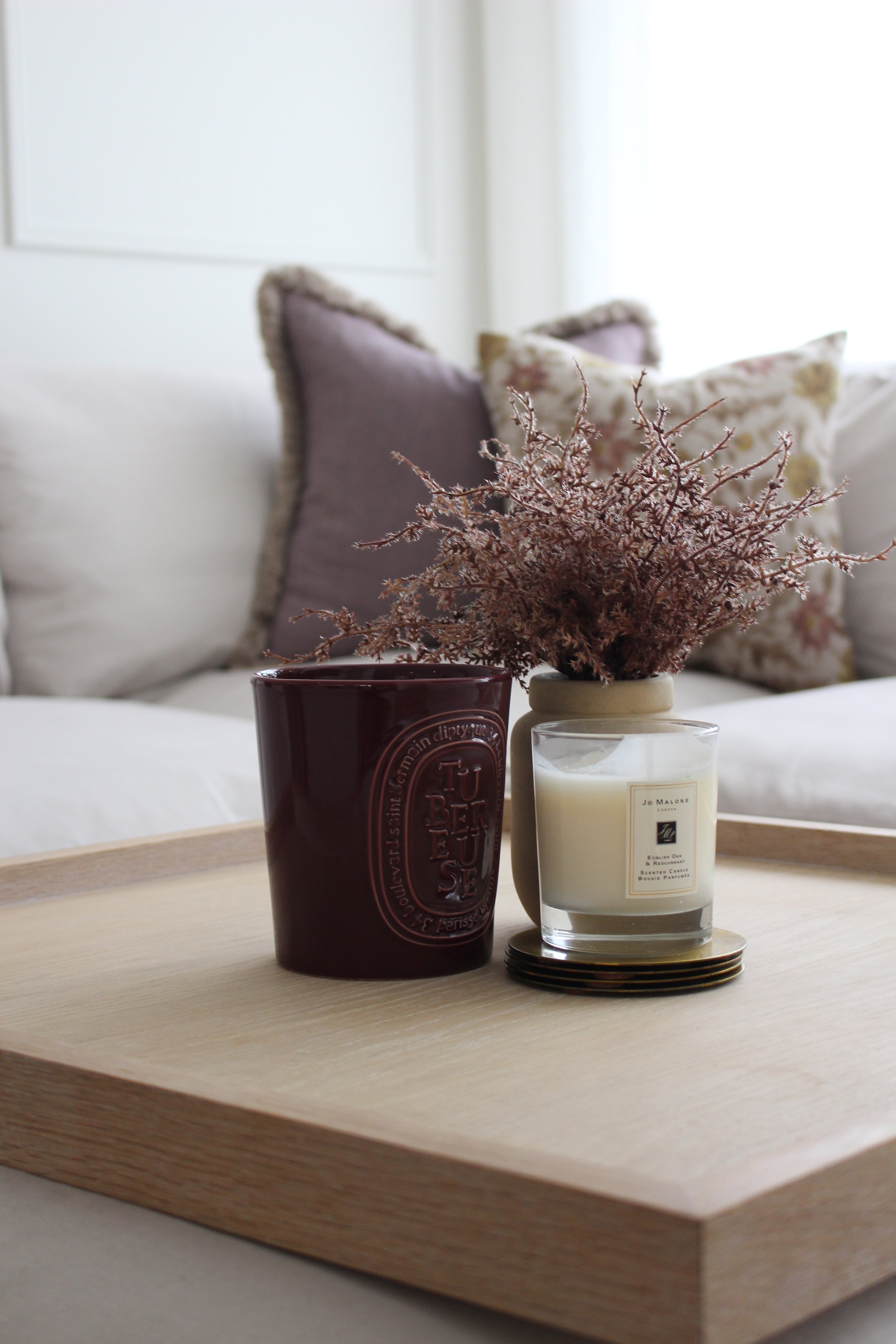 Decorative tray with burgundy dried flowers in a small beige vase, a white candle in a glass container, and a burgundy ceramic candle, all placed on a wooden tray on a coffee table in a cozy living room with burgundy and floral cushions.