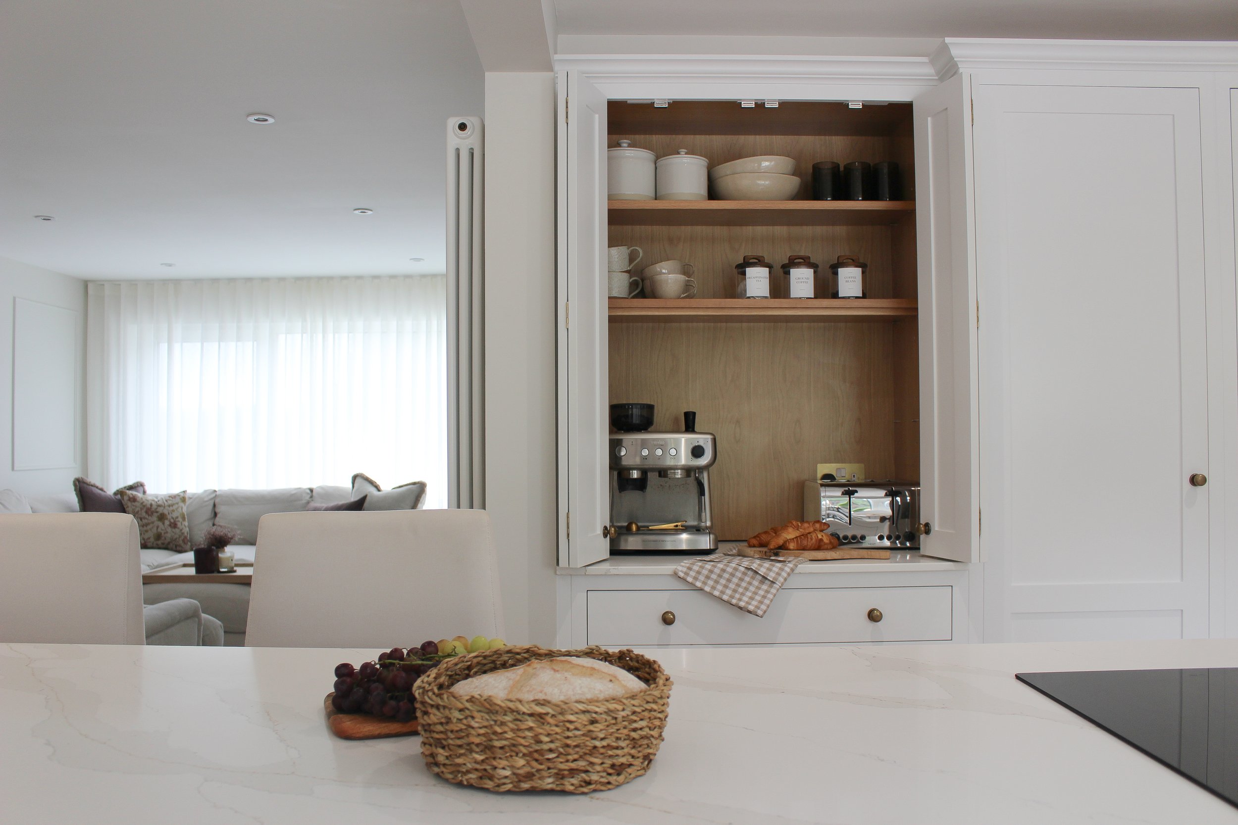 Kitchen with white cabinets, coffee machine, toaster, croissants, grapes, and bread on a white countertop, living room with white sofa and large window with sheer curtains in the background.