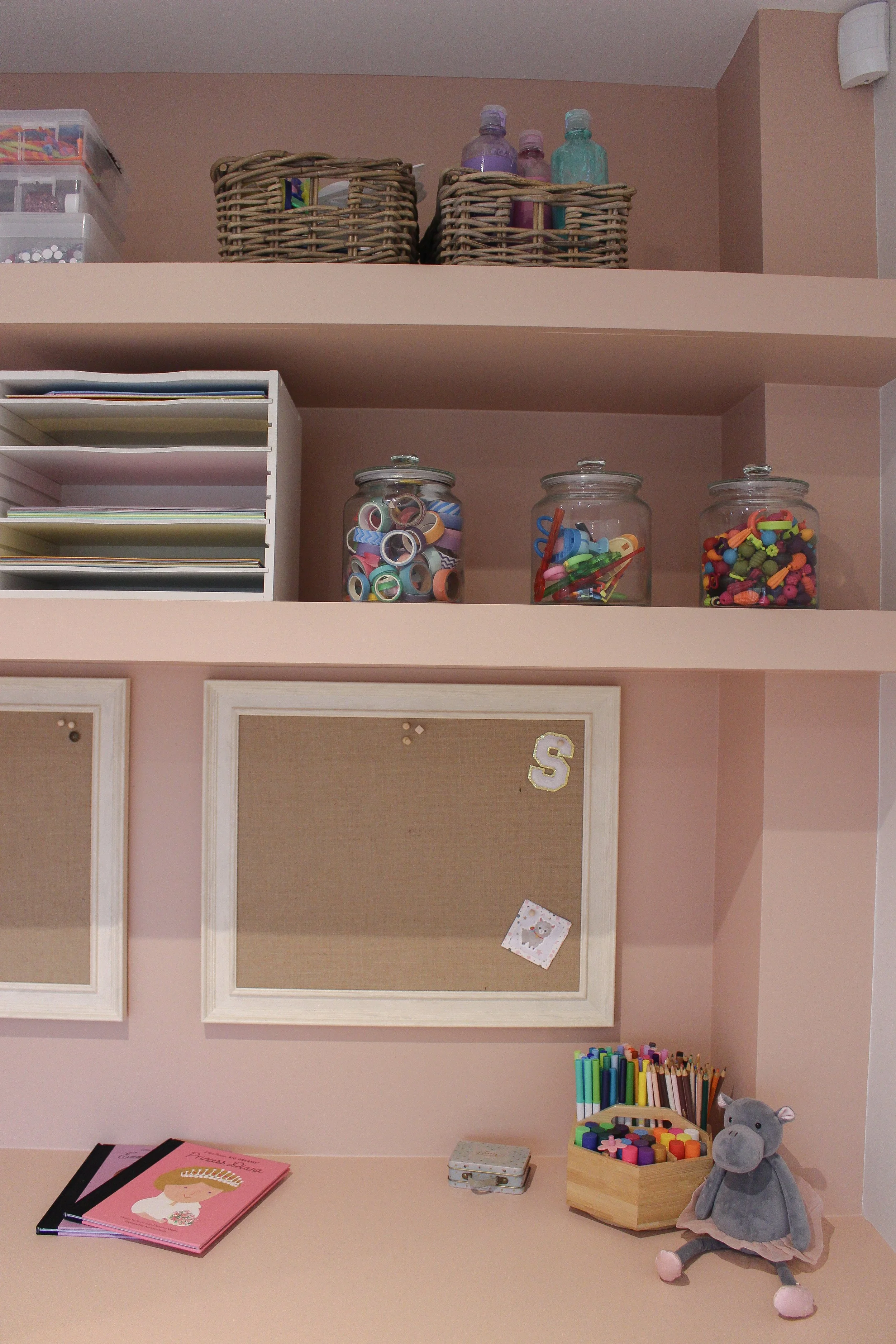 A neatly organised craft or office workspace with shelves holding jars filled with colorful supplies, a corkboard, and a collection of markers and a stuffed toy on the desk. Playroom designed by Yazzmin Lovelle Interiors