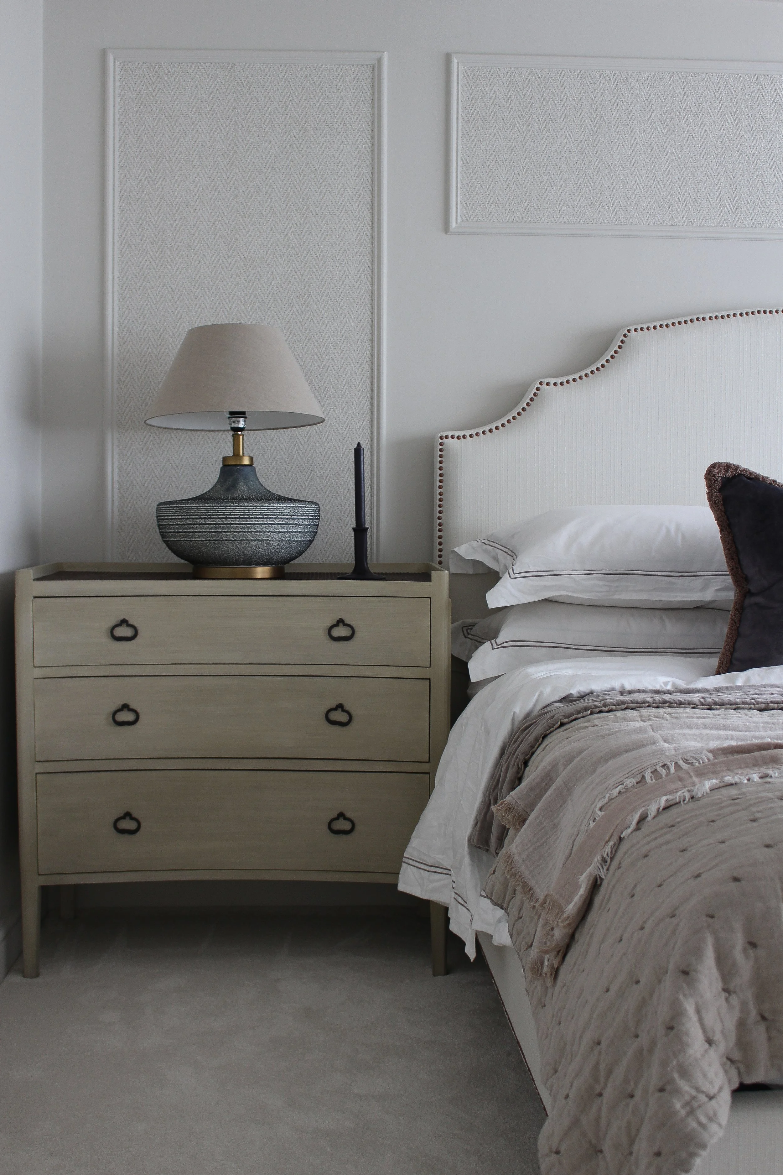 A master bedroom featuring a beige large bedside table with bronze hardware, a textured table lamp, a black candle, a white headboard, and a beige quilted bedspread. A master bedroom designed by Yazzmin Lovelle Interiors