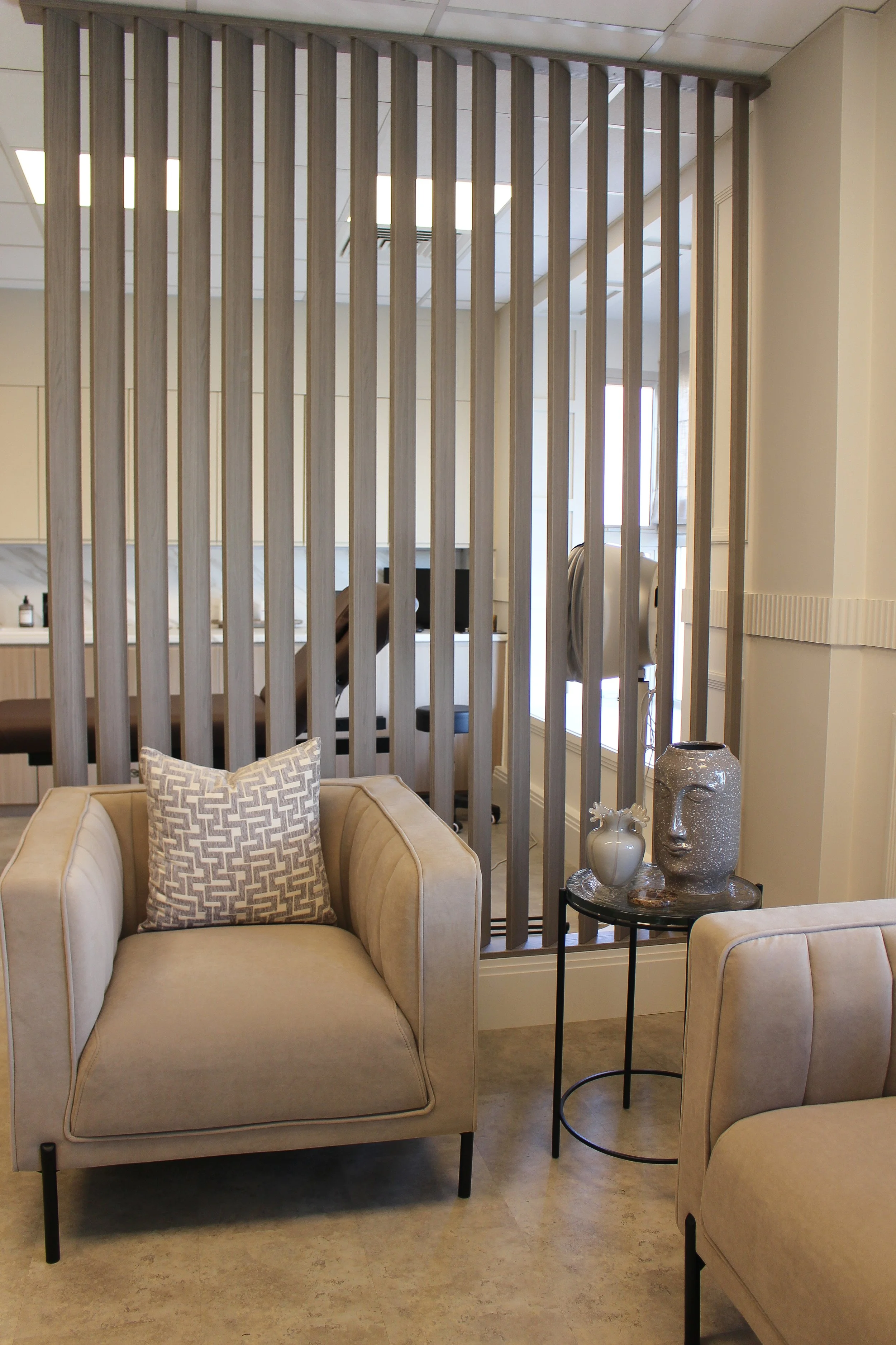 A clinic seating area with two beige armchairs, one with a decorative pillow, a black round side table with vases, and a wooden partition separating it from a treatment area in the background. Clinic designed by Yazzmin Lovelle Interiors