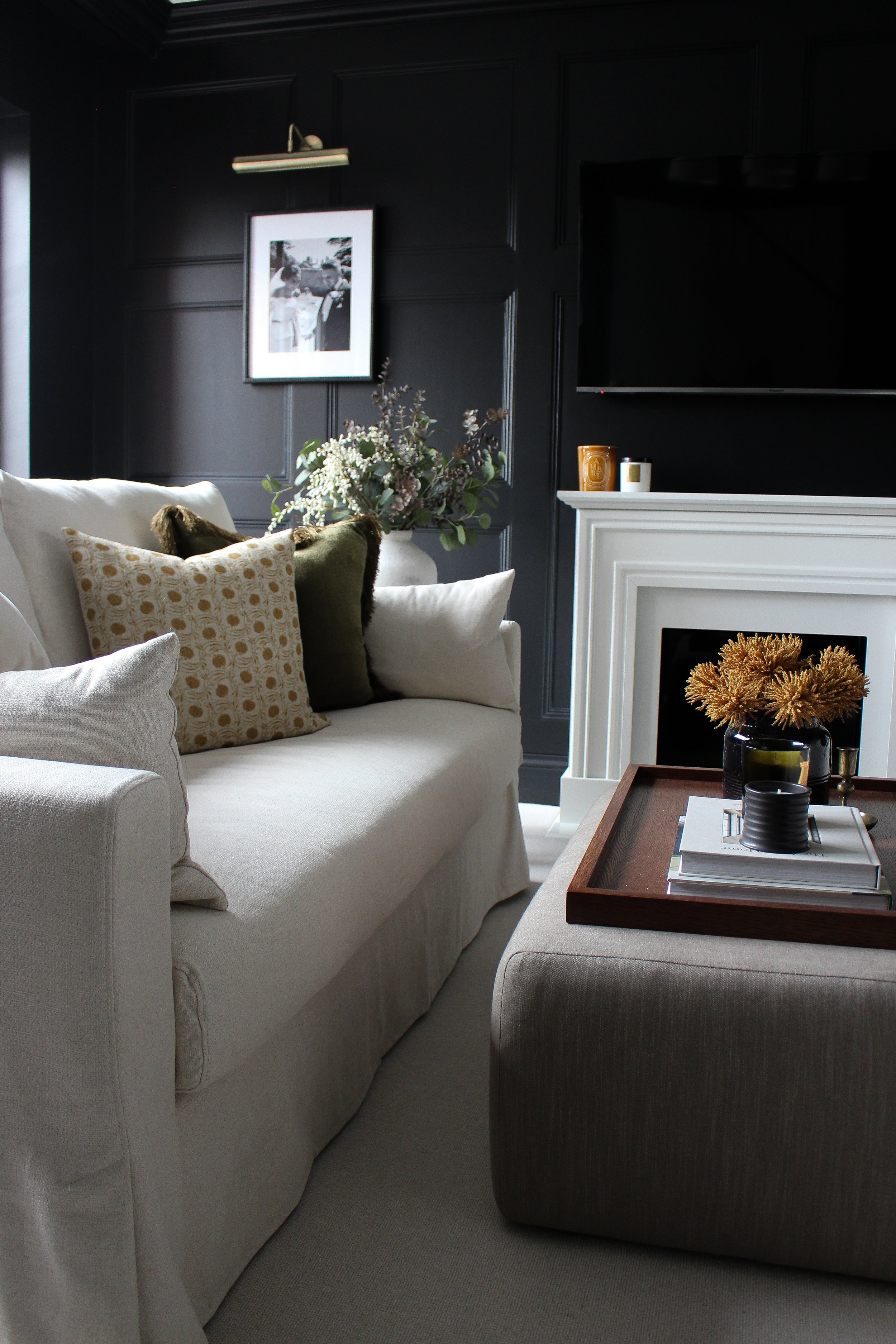 Living room with beige sofa, assorted decorative throw cushions, black panelled wall, white traditional fireplace mantle, floral arrangement, and coffee table with books and vases. Formal lounge designed by Yazzmin Lovelle Interiors.