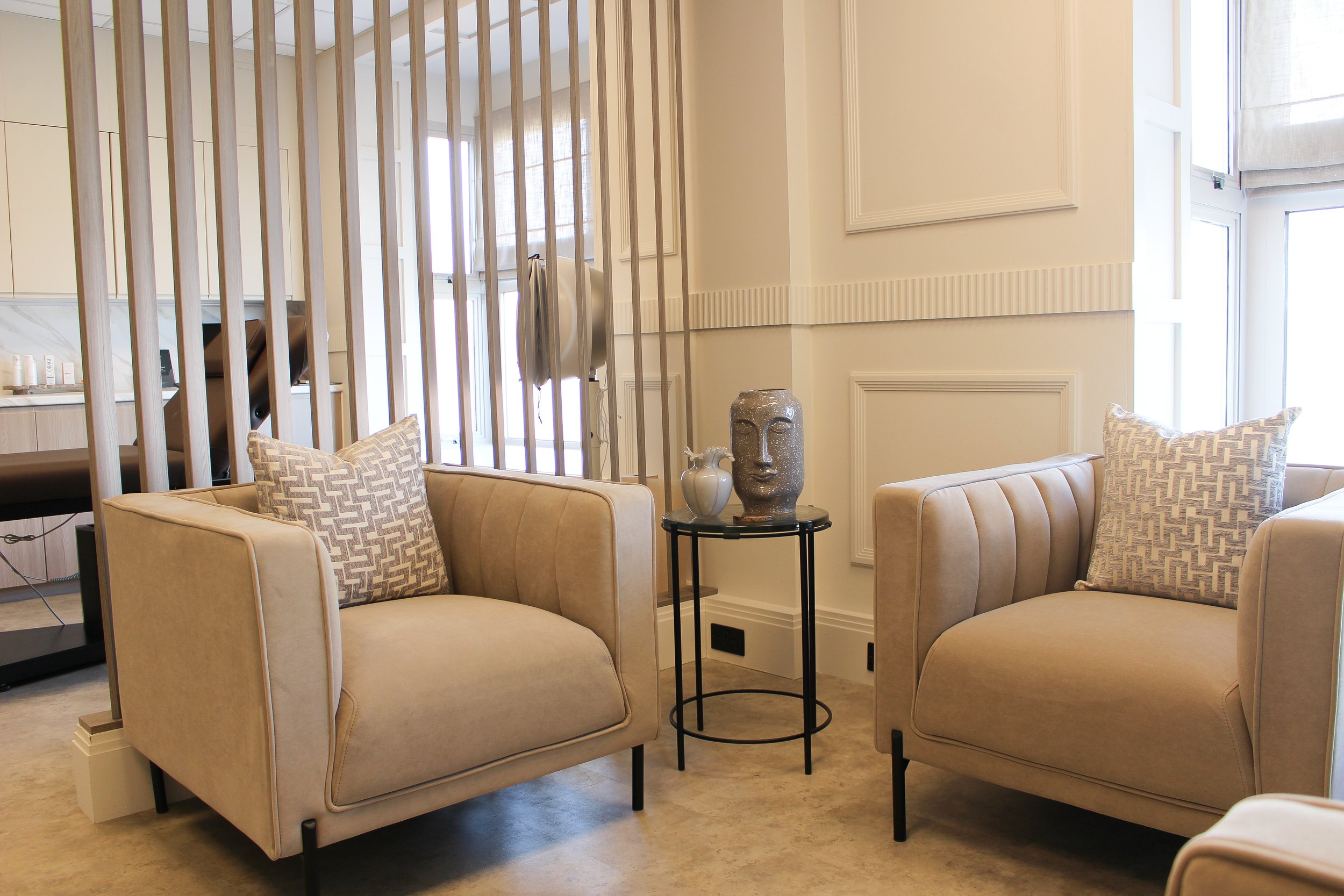 Seating area with two beige armchairs with geometric throw pillows, a small black side table with a decorative face sculpture, and large windows allowing natural light. Clinic designed by Yazzmin Lovelle Interiors