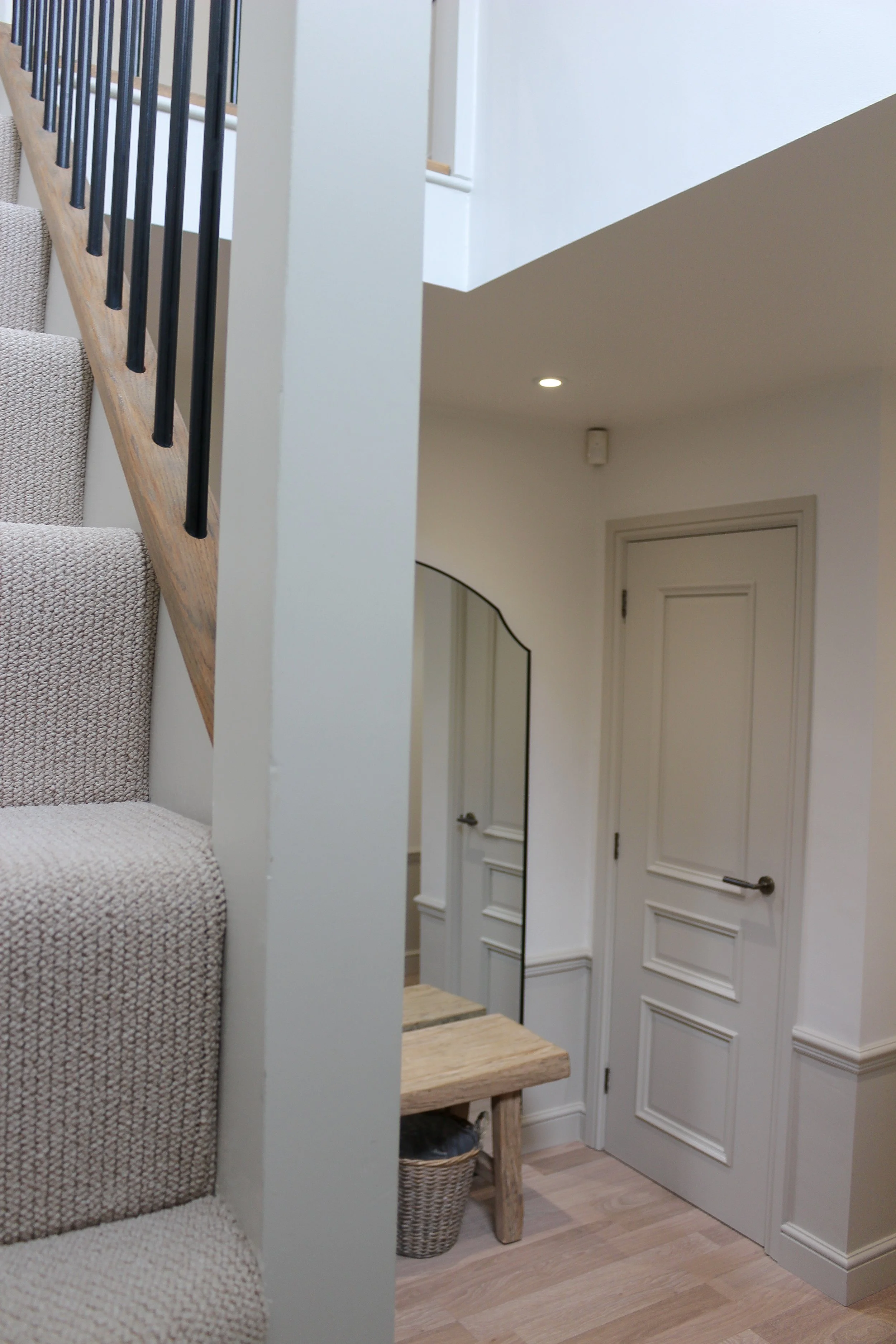 Interior view of a home staircase with beige carpet, a wooden step, and a small wooden bench underneath. A mirror with a black border is visible on the wall near a closed white door with paneling, and baskets under the bench. 