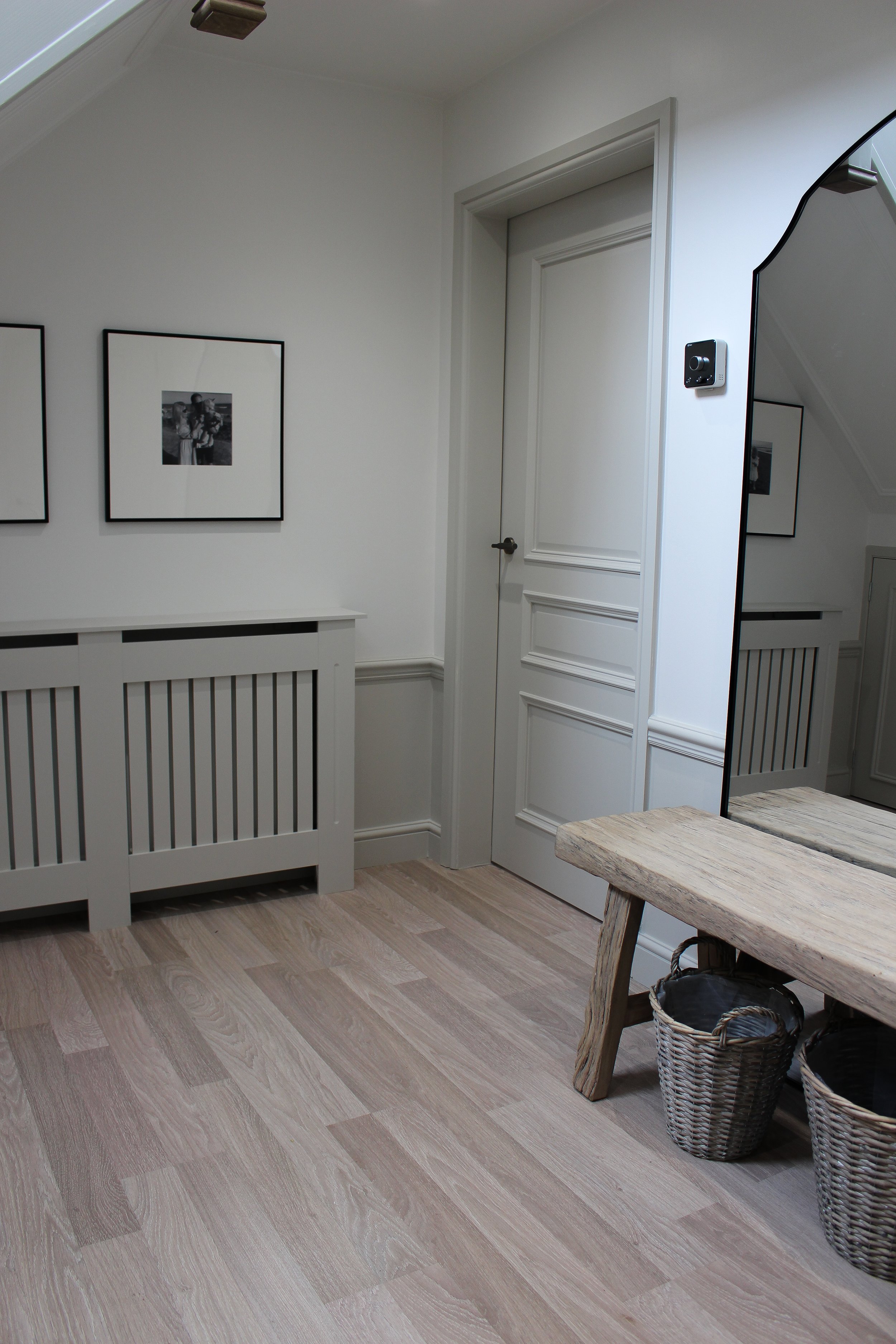 A corner of a hallway with white walls, a wooden floor, a large mirror, a wooden bench, and baskets underneath. Hallway designed by Yazzmin Lovelle Interiors 