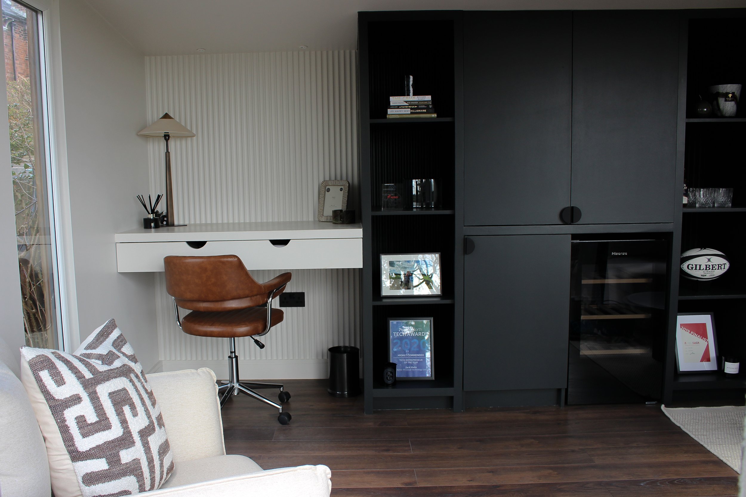 Interior of a modern officr with a white armchair, patterned pillows, a brown leather office chair, a white wall-mounted desk, a black shelving unit with books and framed photos, a small lamp, and a dark wood floor.