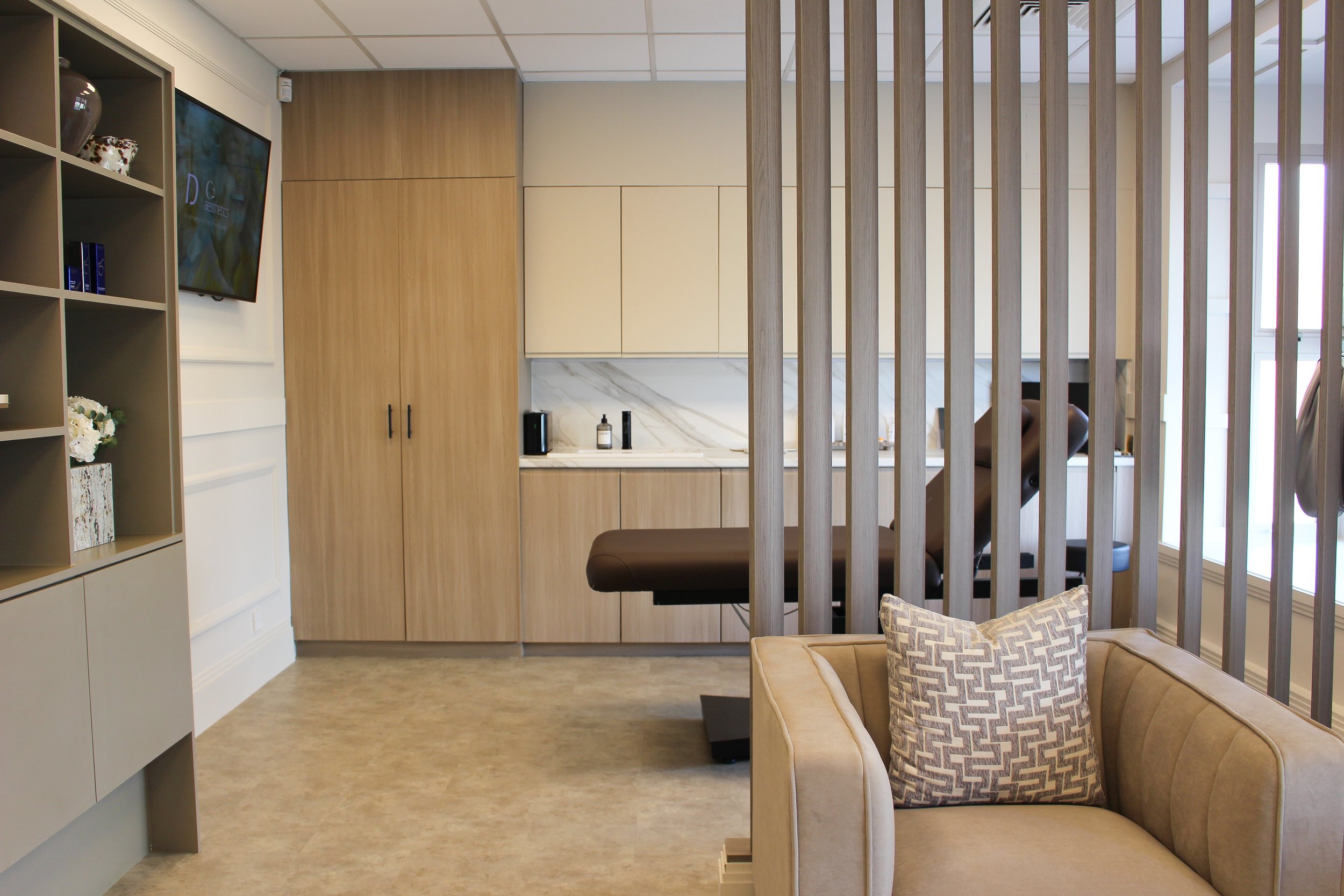 A modern clinic treatment room with a brown treatment chair, wooden storage cabinets, a marble topped counter, a wall mounted television, an armchair with a decorative pillow, and wooden slats dividing the space.