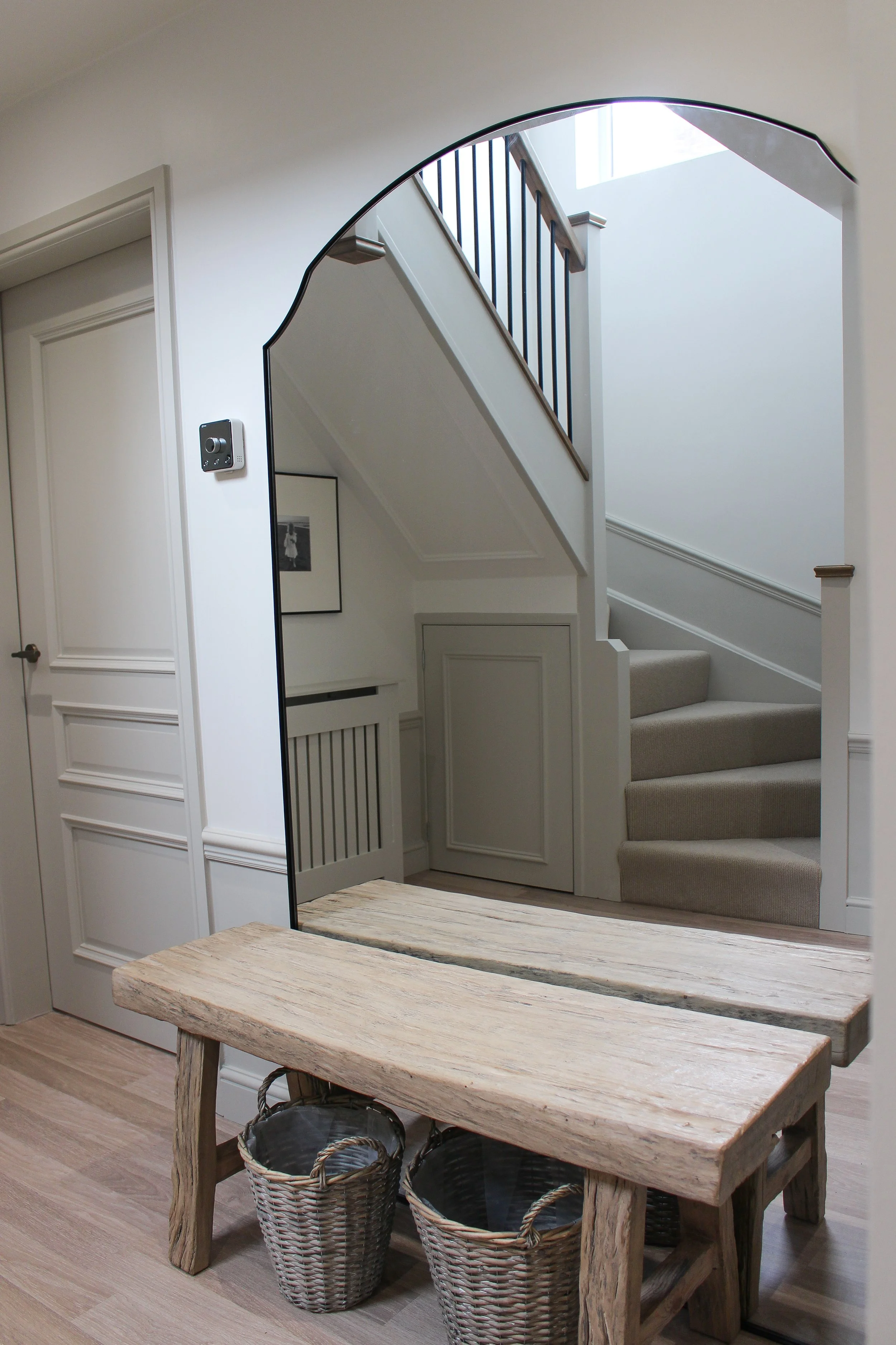 A hallway with a wooden bench and woven baskets in front of a large mirror. Visible stairway with beige carpet and black railing, and a framed black and white photo on the wall. Hallway designed by Yazzmin Lovelle Interiors 