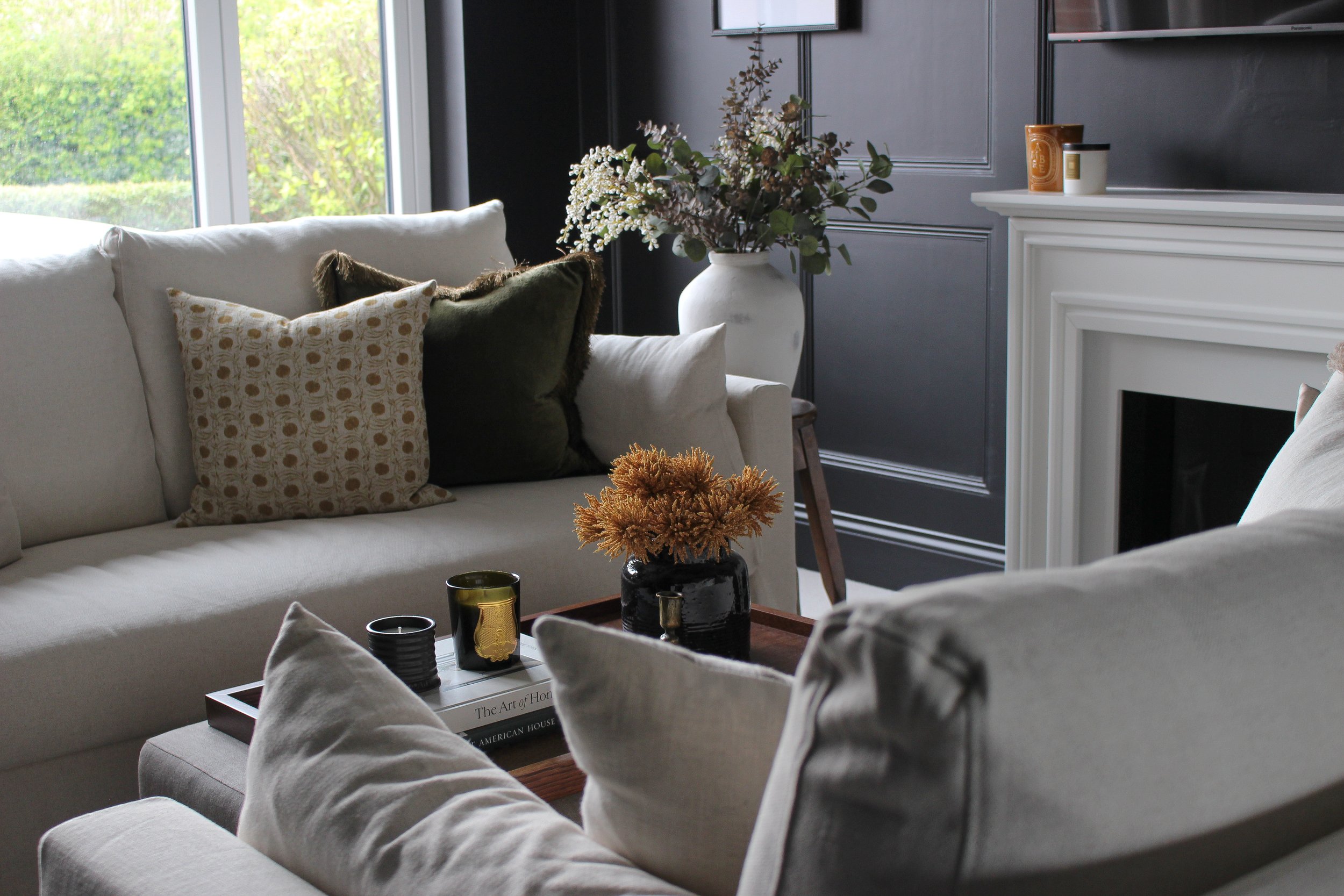 Living room with cream linen sofas decorated with patterned and velvet throw, a wooden tray on a fabric ottoman with books, and a large white stone vase with flowers near a dark wall with a fireplace mantel and a window showing green foliage outside.
